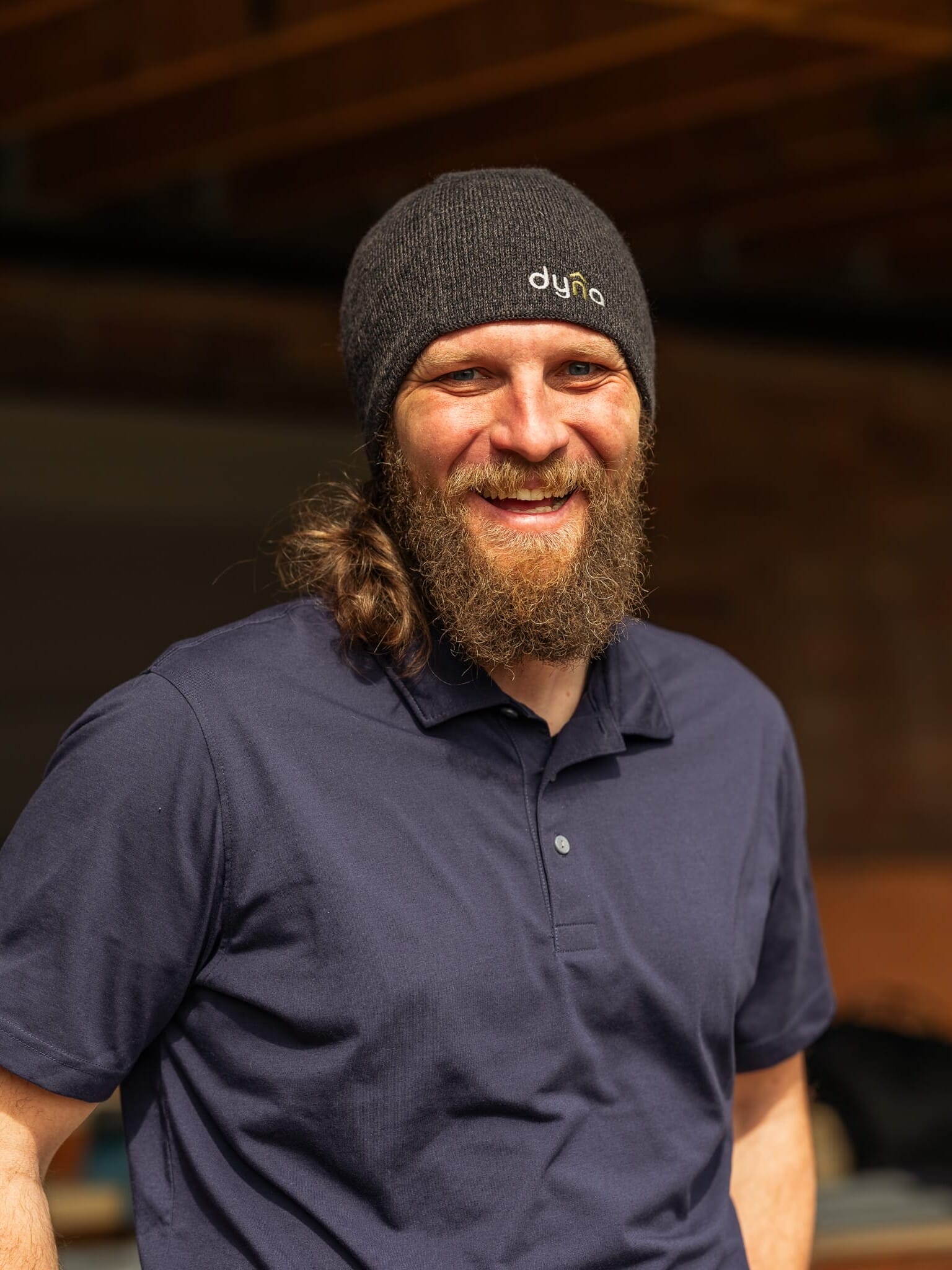 A bearded man with a beanie smiles in front of a building.