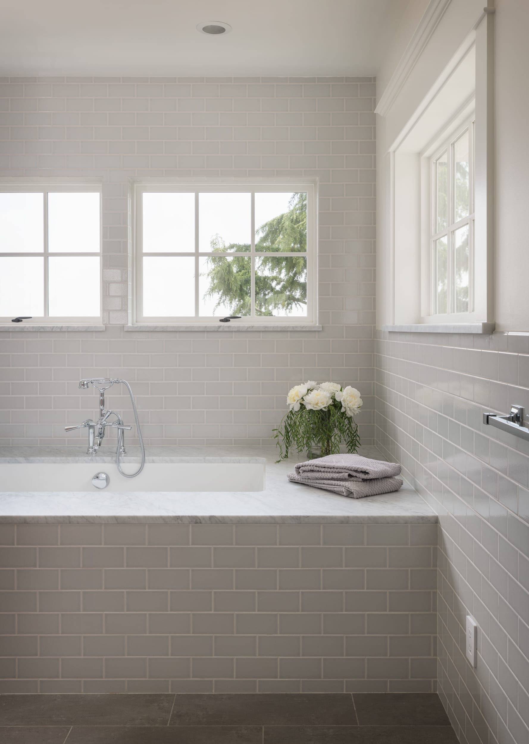 A modern bathroom with white tiles, a tub, and two windows.