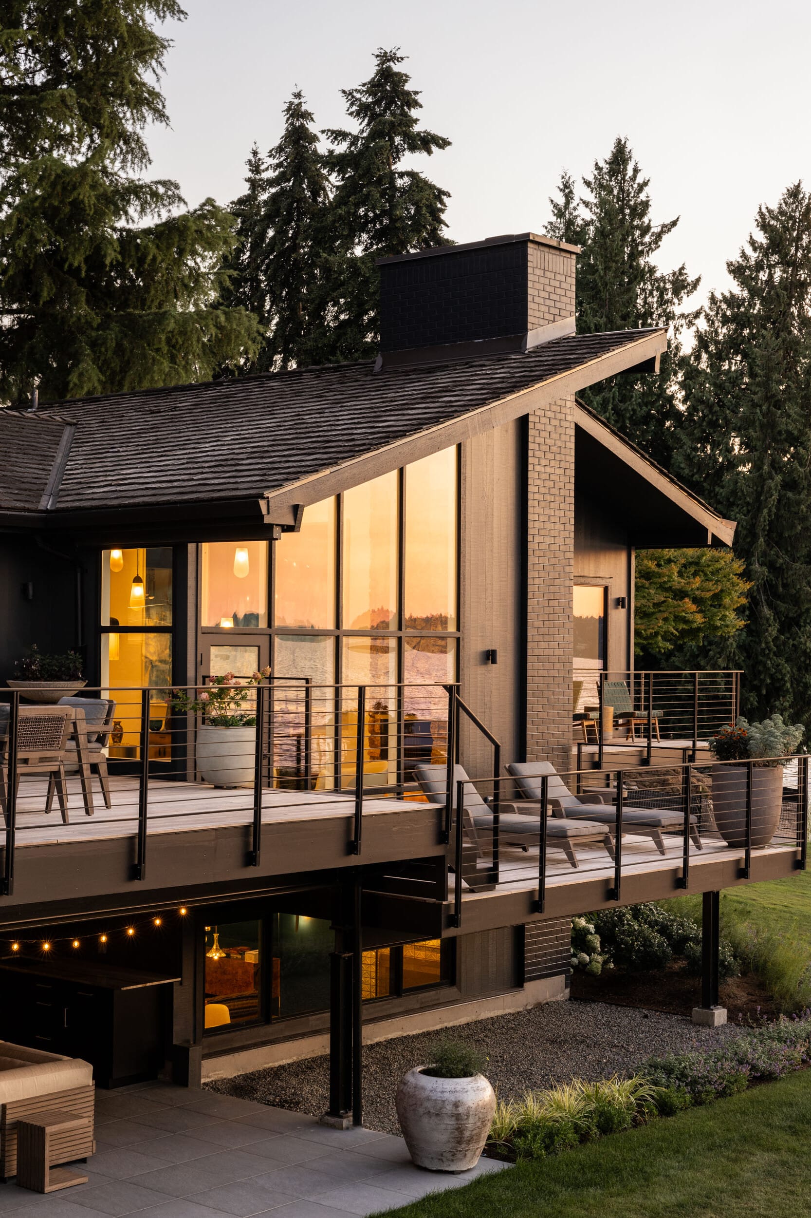 Modern two-story house with large windows and a wraparound deck, surrounded by trees. The interior is warmly lit, and there is patio furniture on the deck.