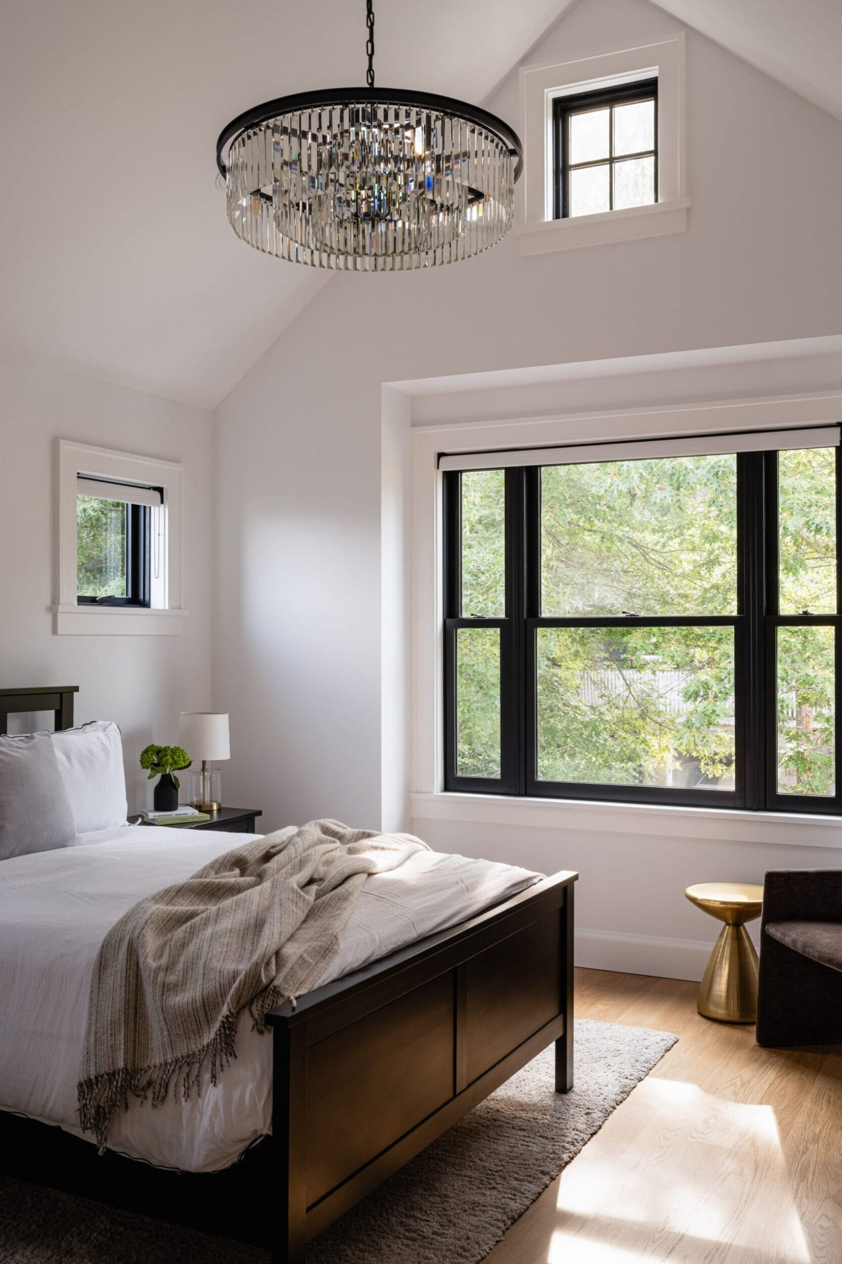 A modern bedroom with a black-framed bed, white bedding, and a beige throw. Large window, armchair, small gold side table, and a crystal chandelier enhance the space. Light wooden floor.