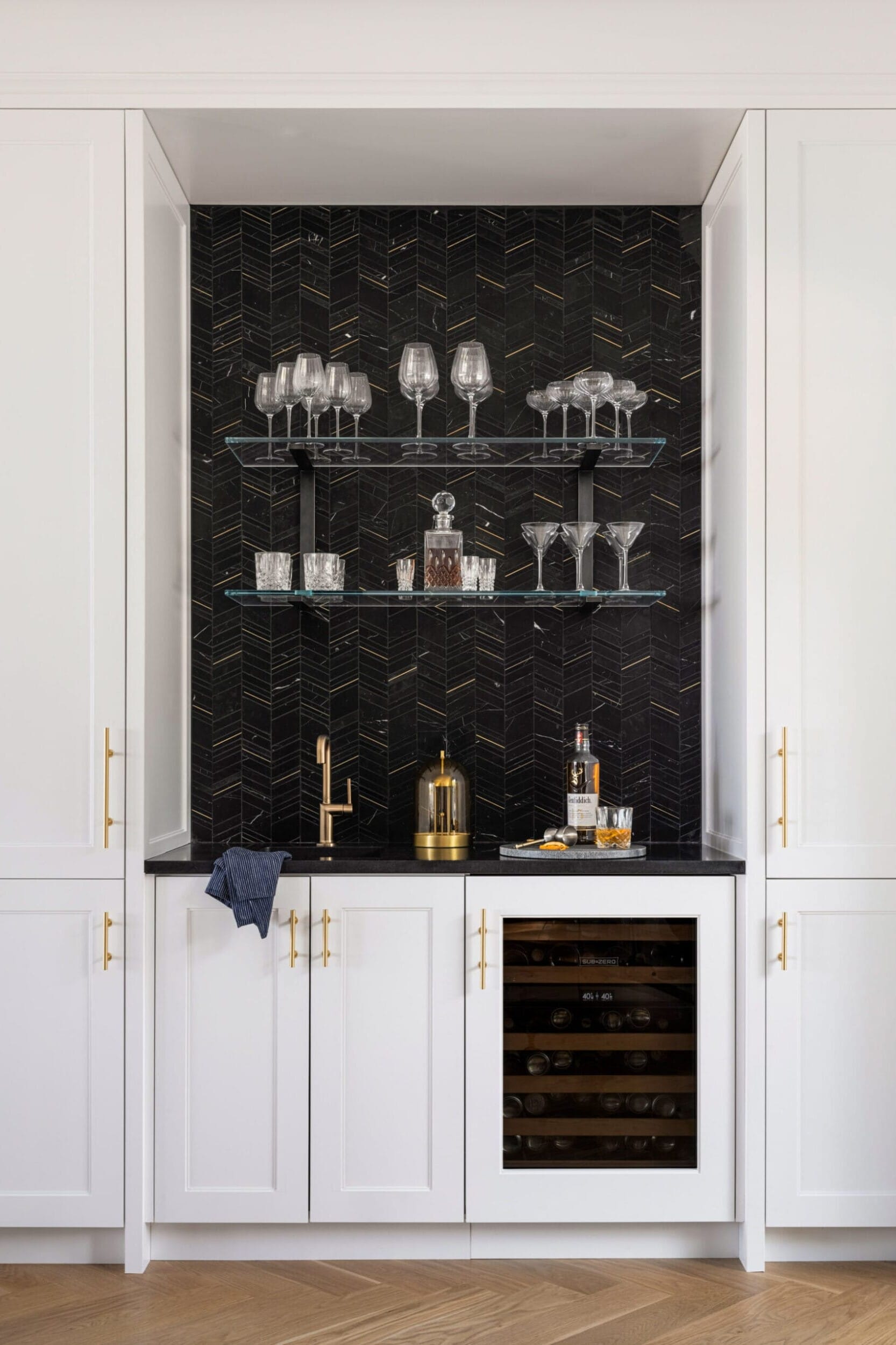 Home bar with a black herringbone backsplash, glass shelves holding glasses, a countertop with a few bar accessories, and a wine fridge with white cabinetry on each side.