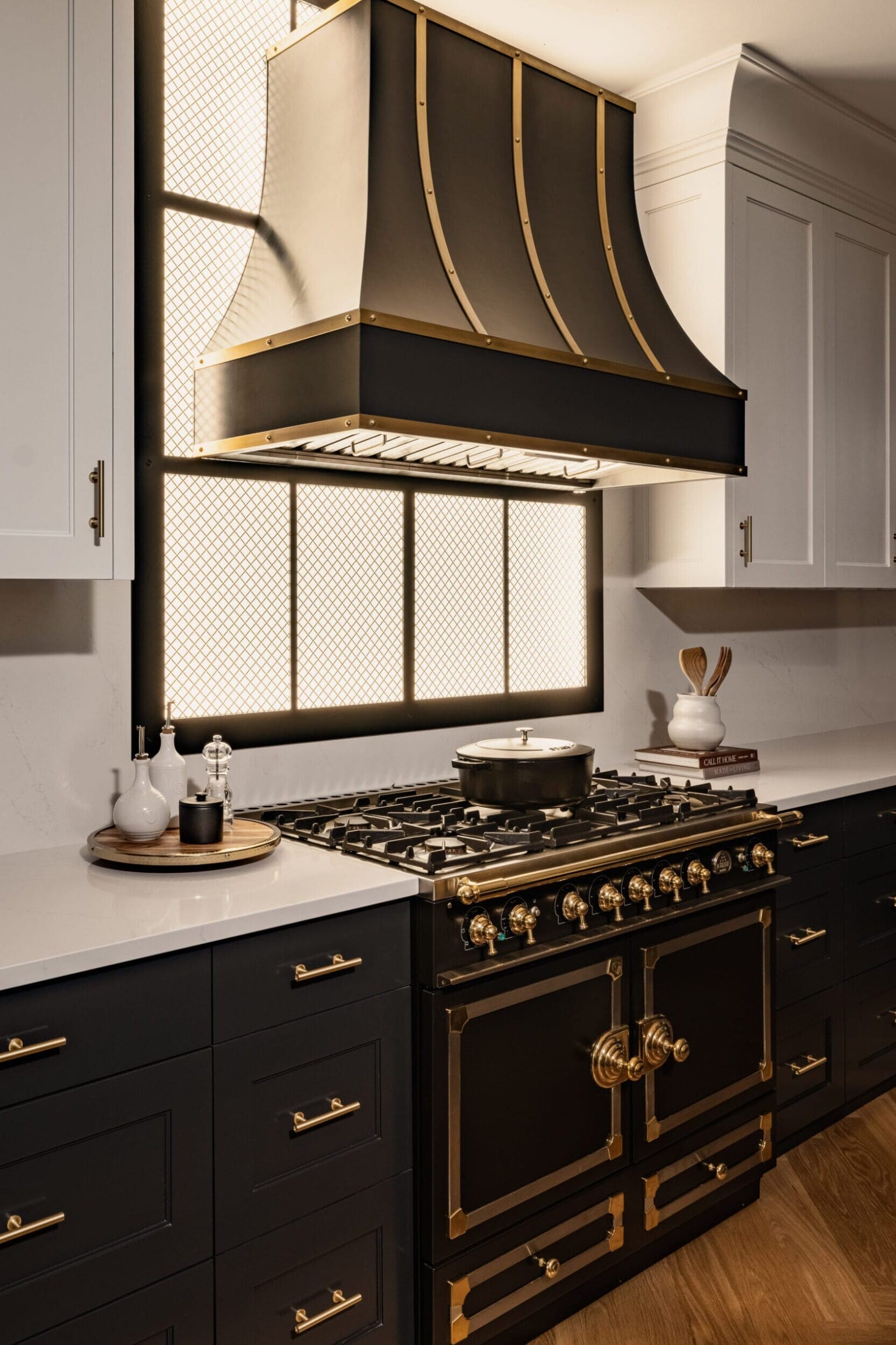 Modern kitchen with a black and gold stove and range hood, white upper cabinets, dark lower cabinets, and wooden floor. A large window provides natural light.