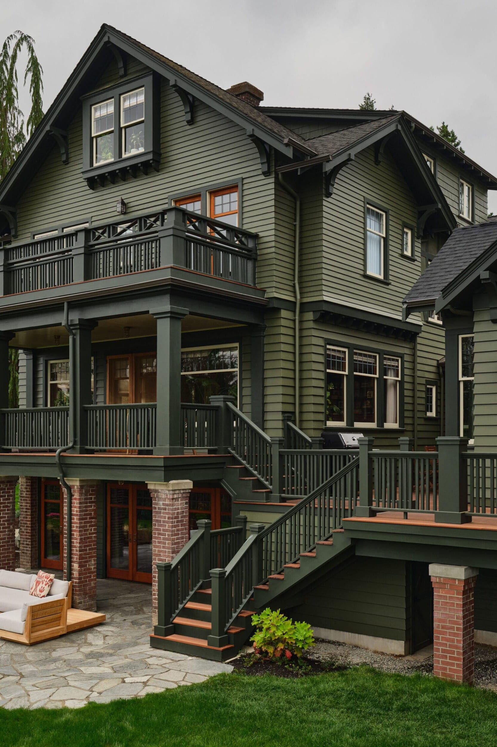 A large green house with multiple balconies and a patio area. It has white-trimmed windows, a staircase leading to the yard, and outdoor seating on stone flooring.