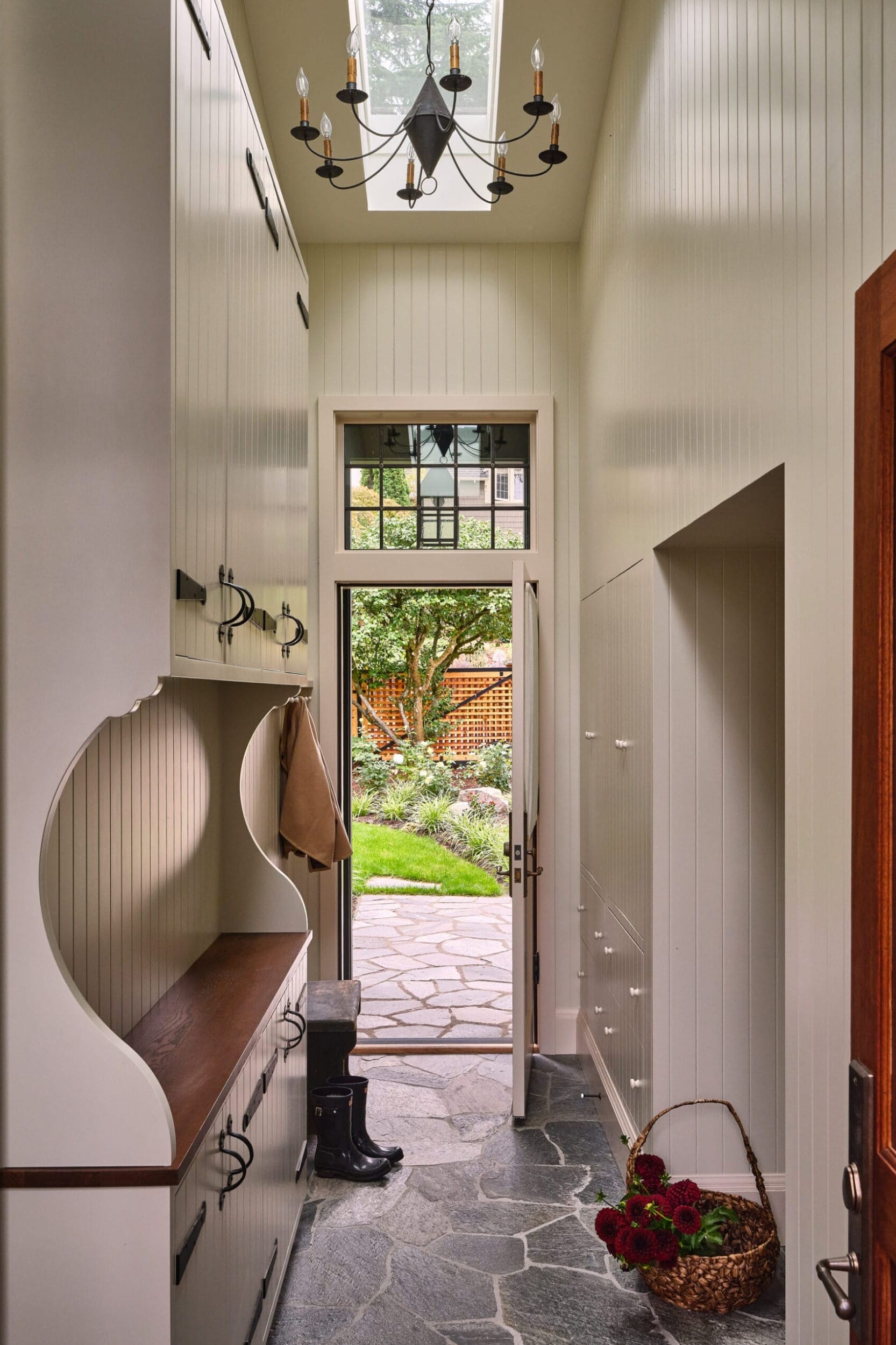 Narrow mudroom with white cabinets, stone floor, chandelier, and open door leading to a garden. Basket with red flowers and black boots in the room.