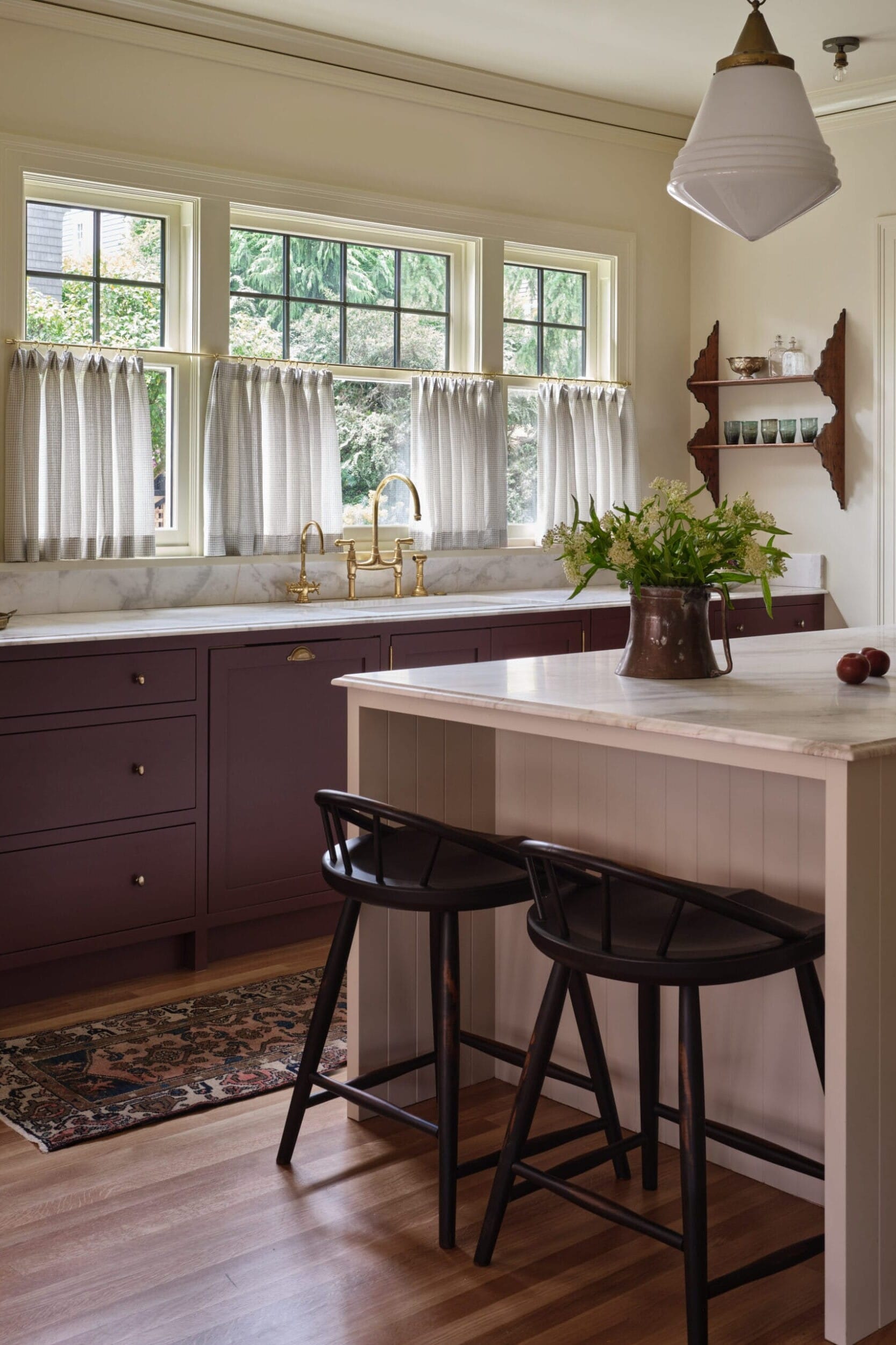 A kitchen with a marble countertop, dark cabinets, a vase with green leaves, two black chairs, and large windows with curtains overlooking trees.