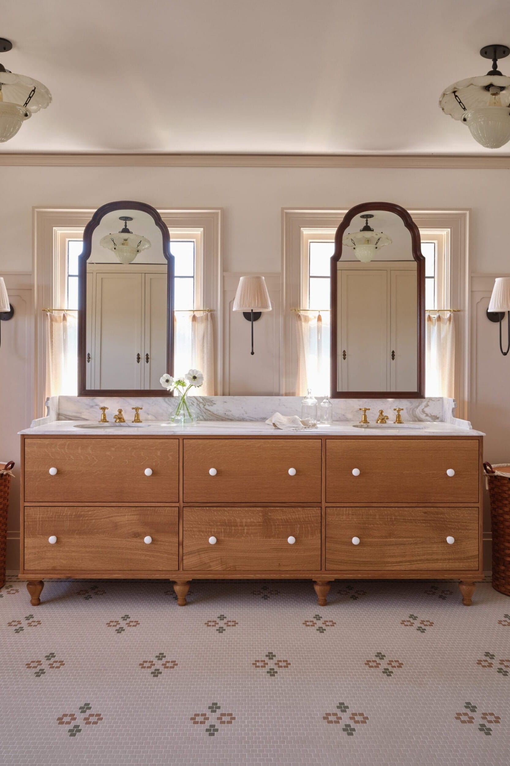 A bathroom features a wooden double vanity with two mirrors, brass faucets, flowers, and wicker baskets. There are wall sconces and two windows above the vanity. The floor has a patterned tile design.