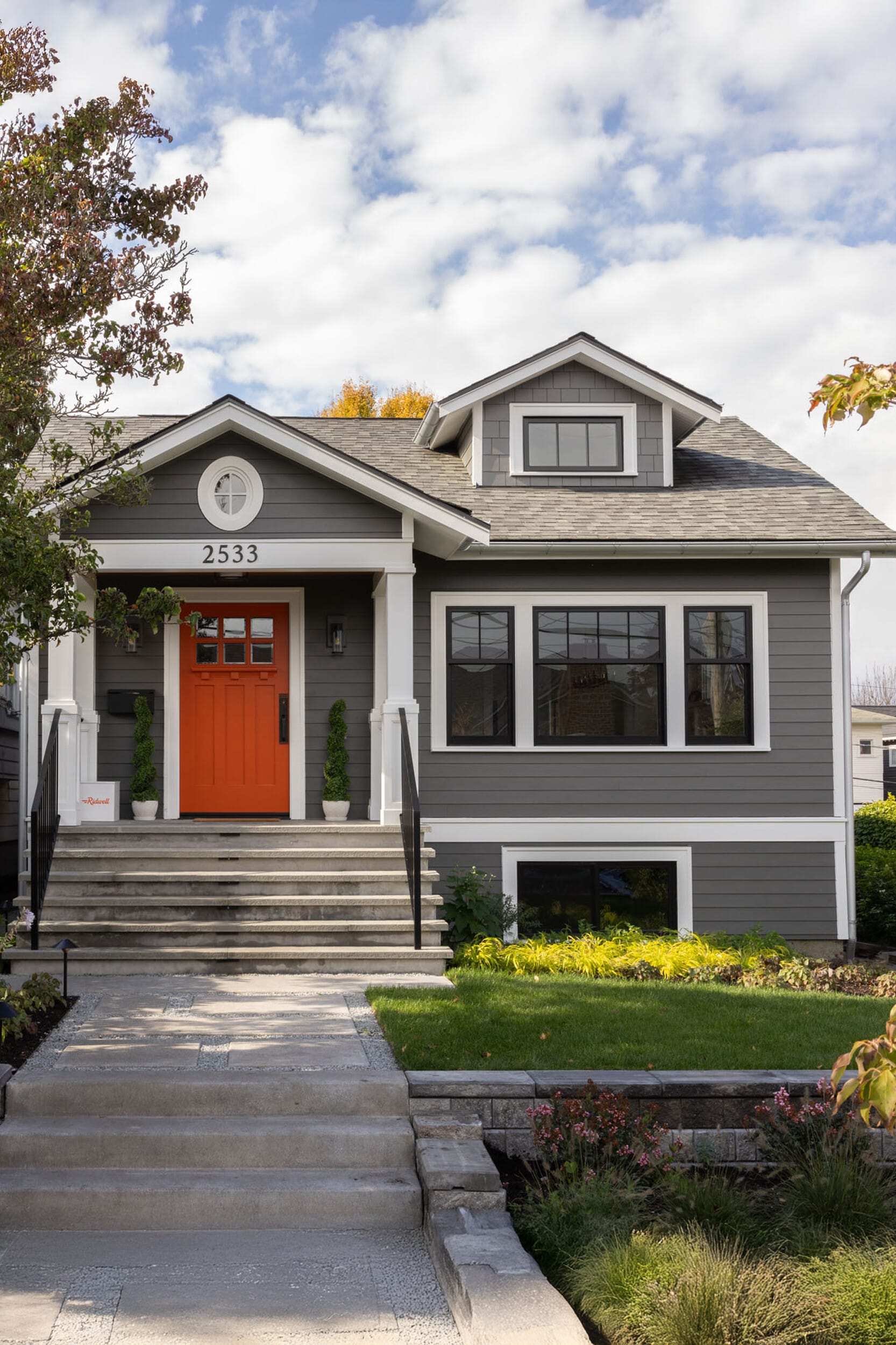 Gray two-story house with an orange front door and the number 2533. Steps lead up to the entrance, surrounded by shrubs and a small lawn. Cloudy sky overhead.