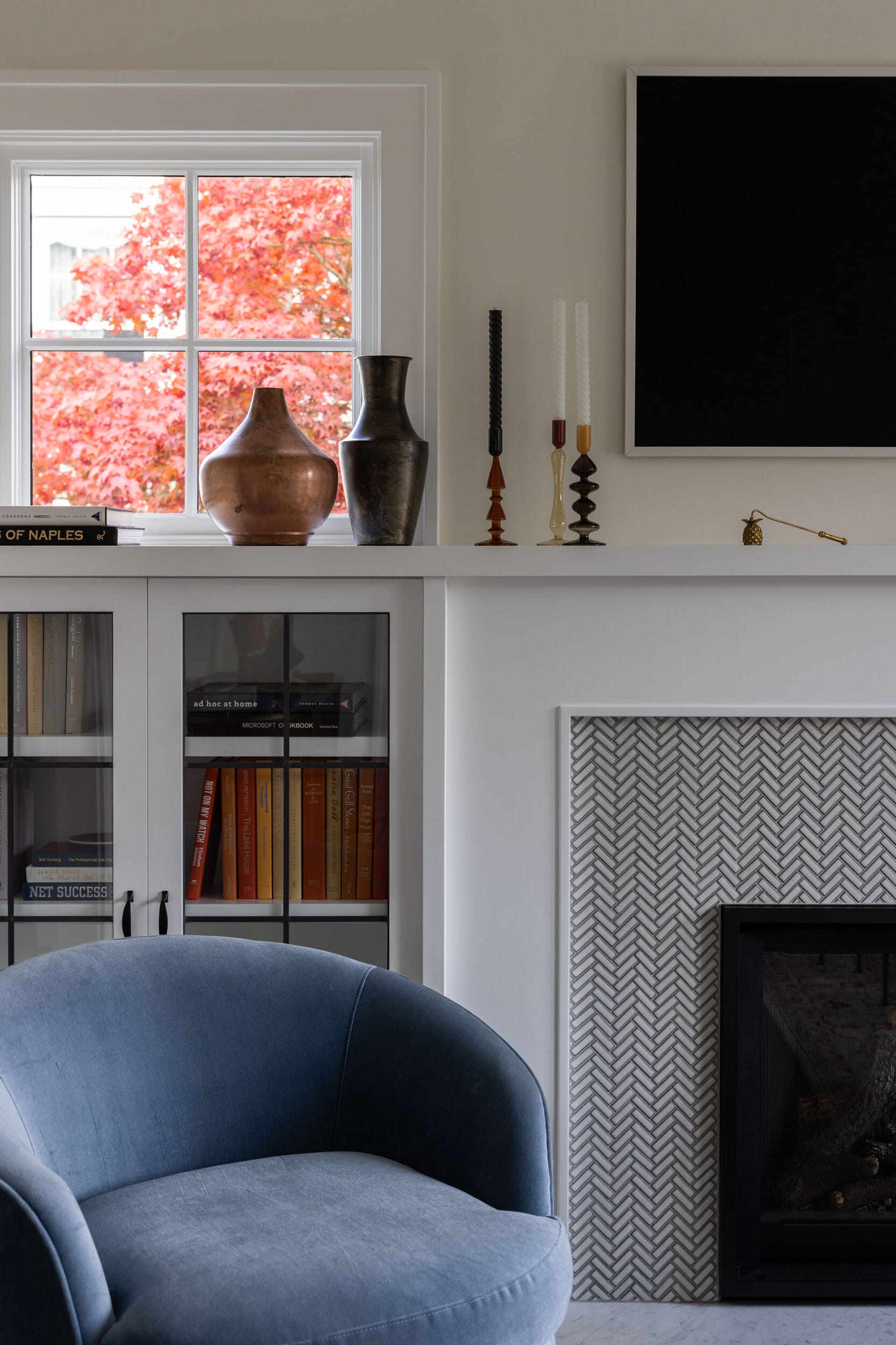 Cozy living room with a blue armchair, a white fireplace mantel featuring vases and candles, a black-framed TV, and a window showcasing red autumn leaves. Bookshelf with books below.