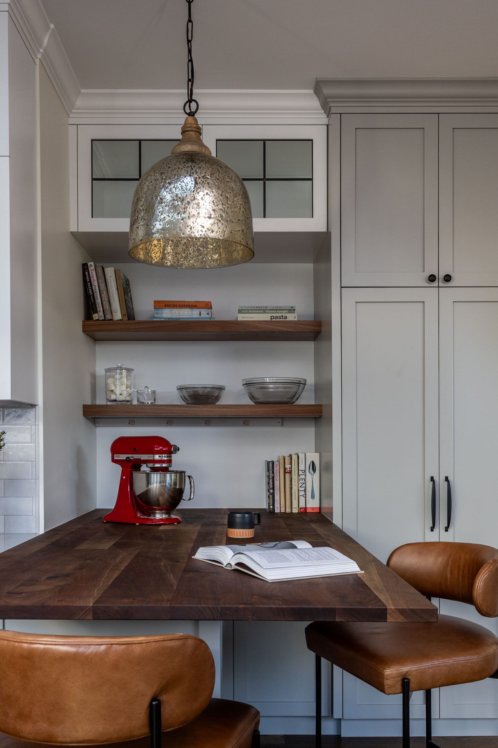 Kitchen with a wooden island, red mixer, open book, two leather chairs, metal pendant light, shelves holding books and bowls, and light gray cabinets in the background.