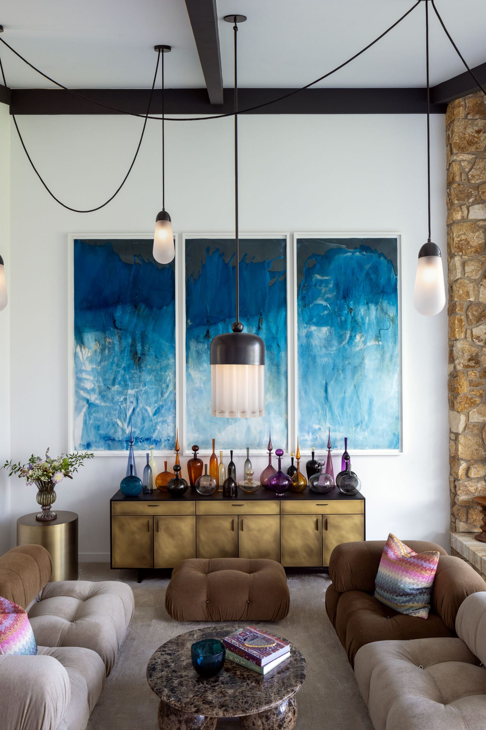 Modern living room with pendant lights, tufted sofas, a stone fireplace, and abstract blue artwork. A sideboard displays colorful glass bottles.