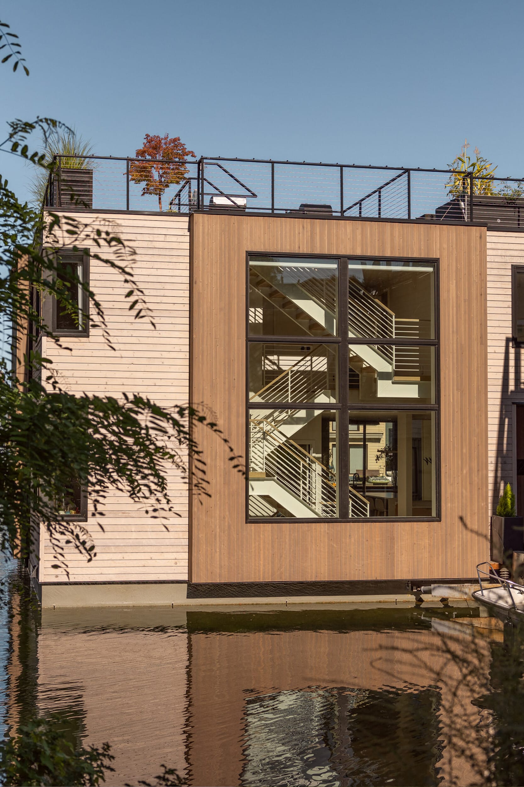 A modern wooden house with large windows sits by the water, reflecting its exterior. A staircase is visible through the window.