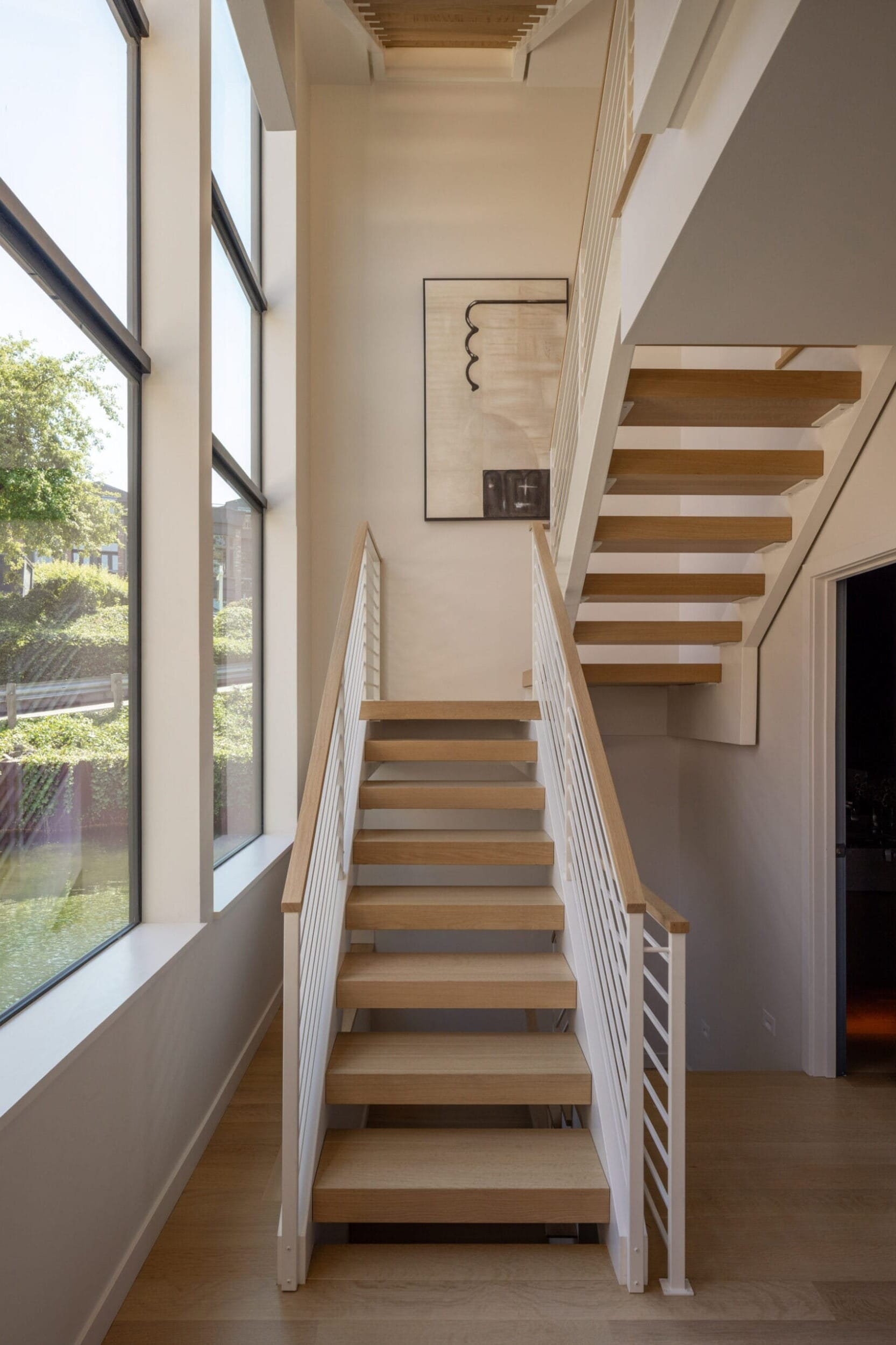 A modern interior staircase with wooden steps and white railings, large windows, and an abstract painting on the wall.