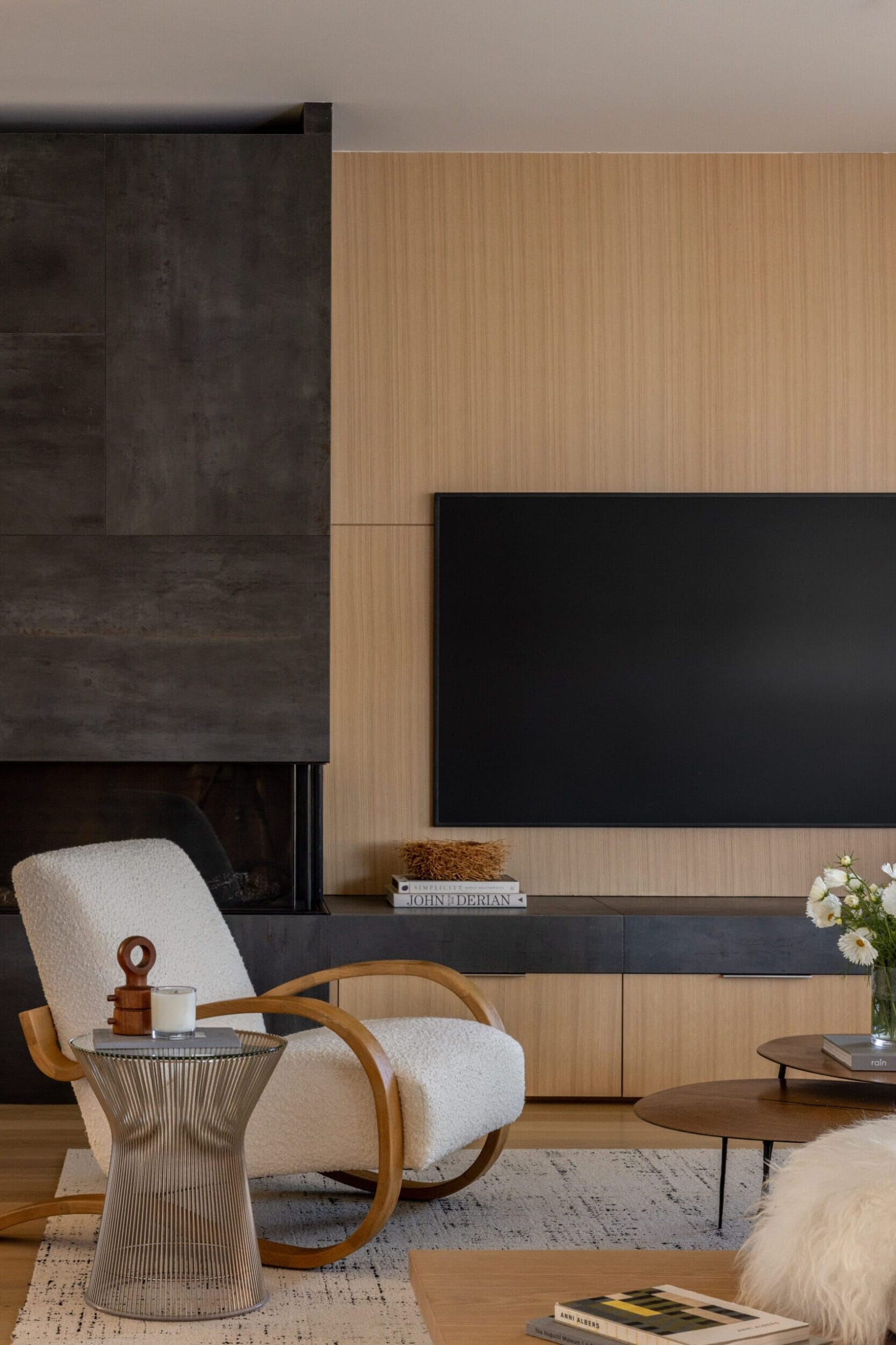 A modern living room with a wooden accent wall, a mounted flat-screen TV, a white rocking chair, a glass side table, books, and a wooden coffee table with white flowers.