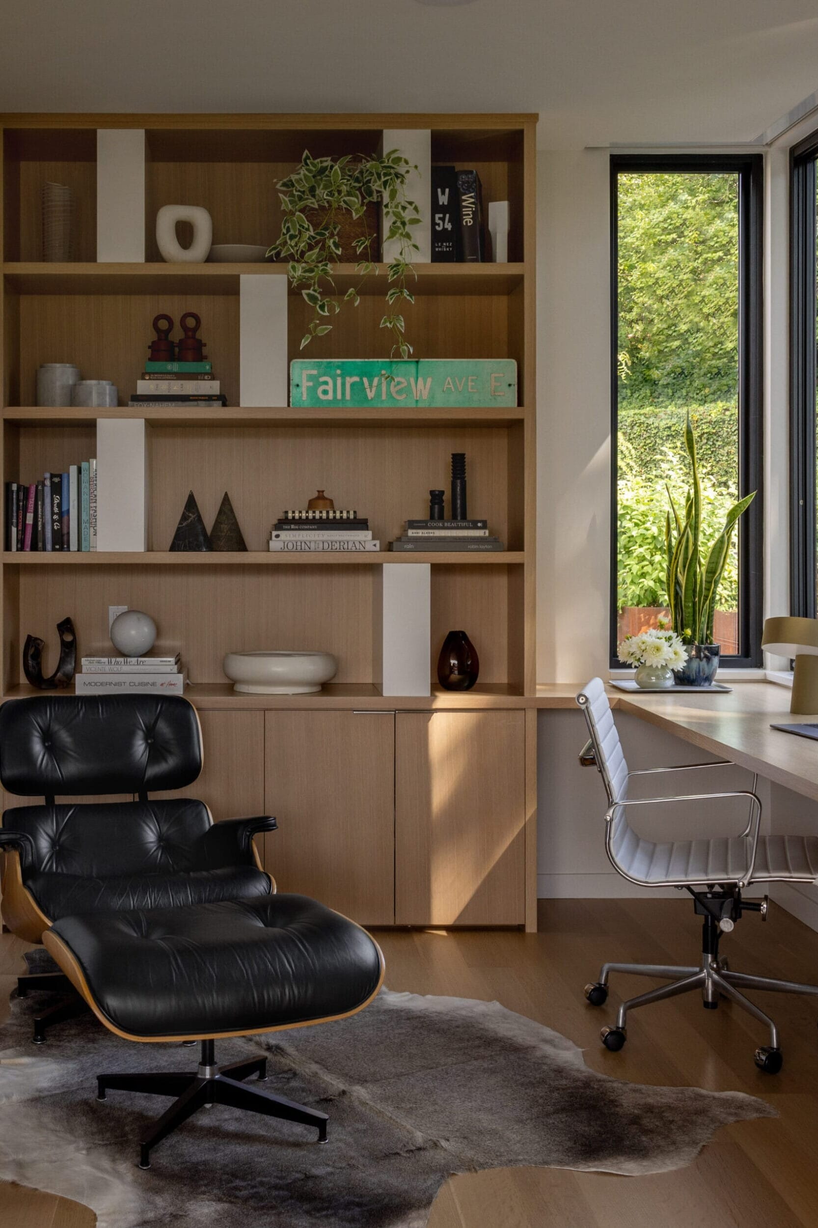 Modern home office with a lounge chair, cowhide rug, wooden shelves with decor, a desk with a white chair, large windows, and a potted plant.