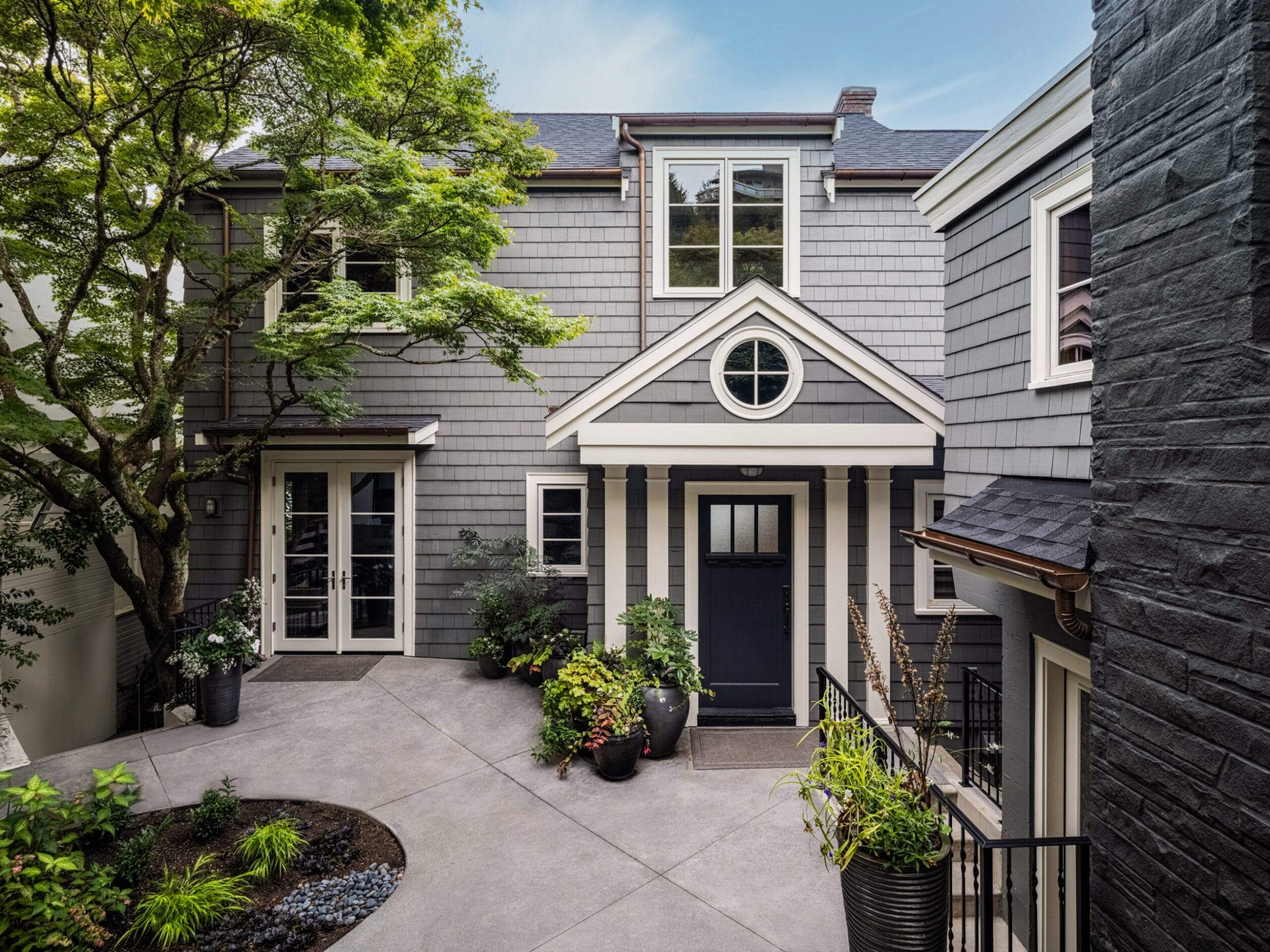 A two-story gray house with a black front door, surrounded by potted plants, and featuring a round window above the entryway. A concrete path leads to the entrance.