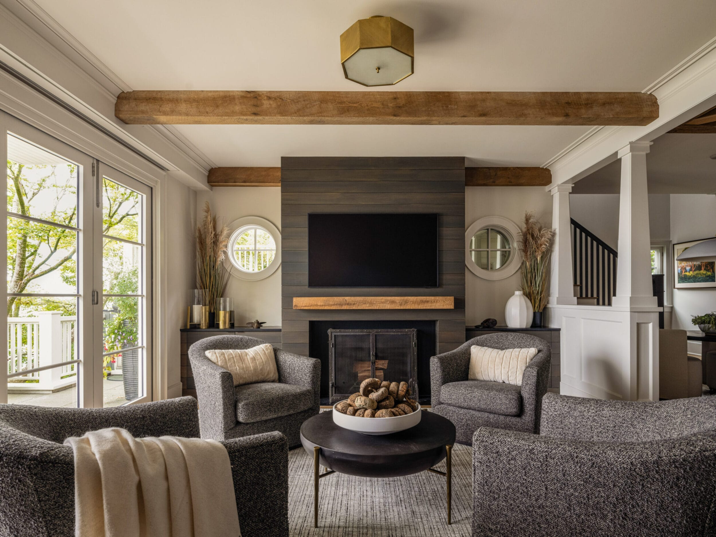 A cozy living room with four gray armchairs around a circular table, a TV mounted above a fireplace, wood beams on the ceiling, and large windows letting in natural light.