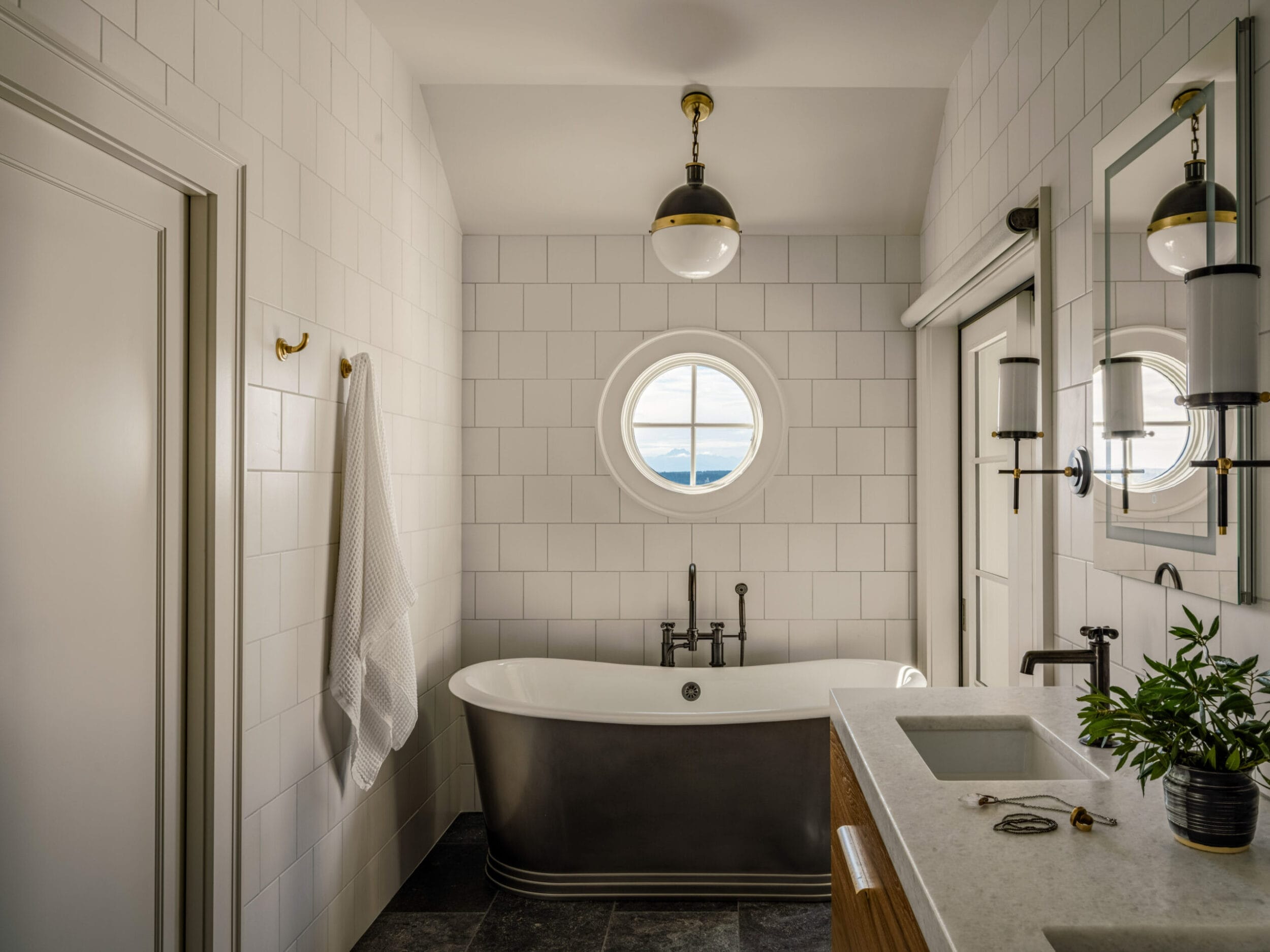 Modern bathroom with a freestanding tub, circular window, and white tiled walls. A towel hangs on a hook, keys are on the counter, and a potted plant sits beside the sink.