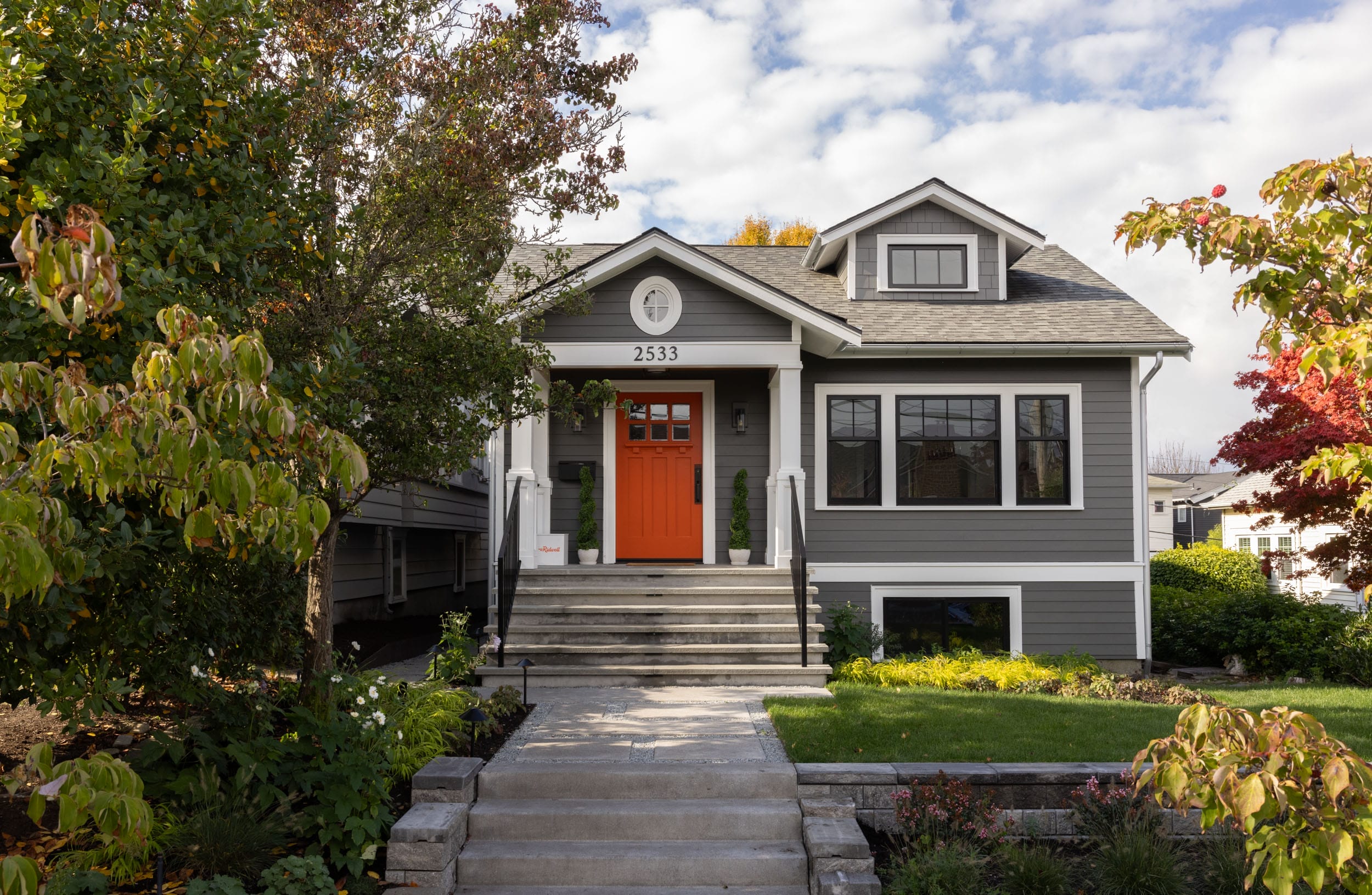 Grey house with an orange front door and stairs, surrounded by trees and shrubs.