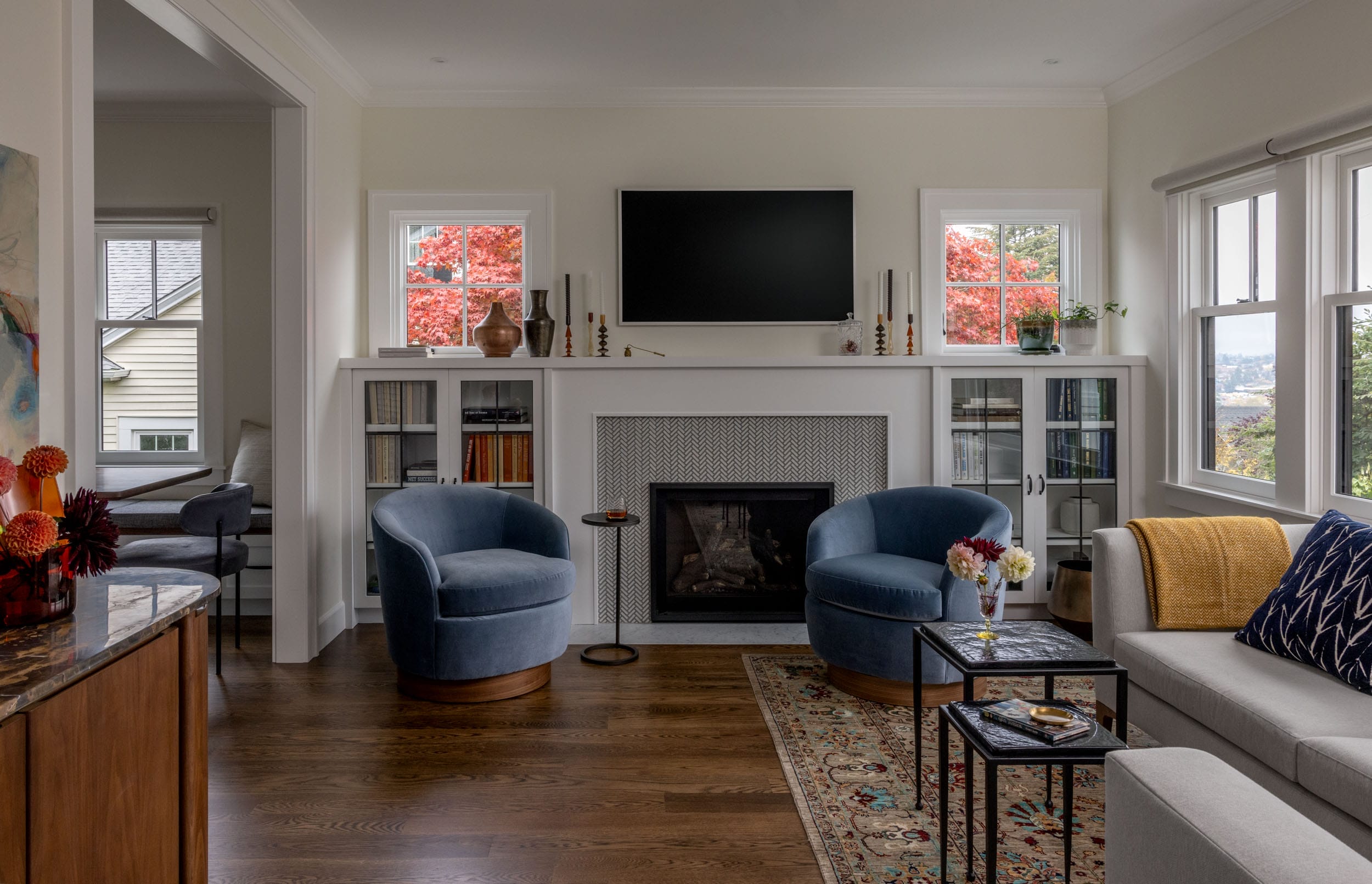 A cozy living room with blue armchairs, a gray sofa, a fireplace, and a TV. Large windows reveal red autumn trees outside. Shelves and a rug complete the decor.