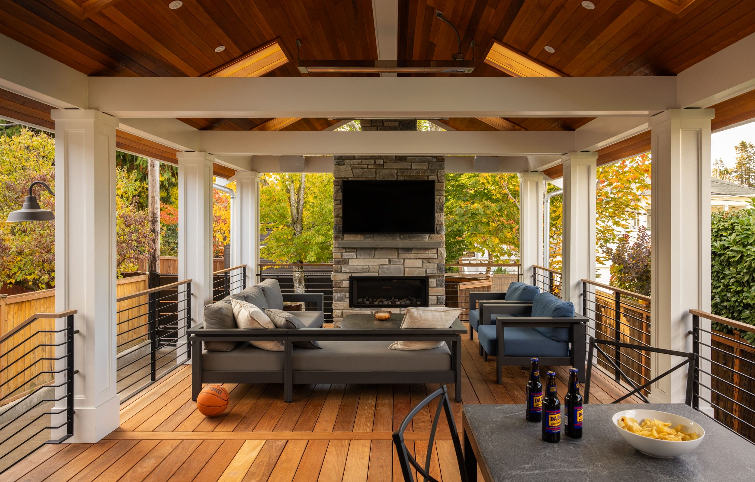 Outdoor seating area with wooden flooring, a stone fireplace, wall-mounted TV, and sofas. A basketball sits on the floor. Bottles and a bowl of chips are on a table in the foreground.