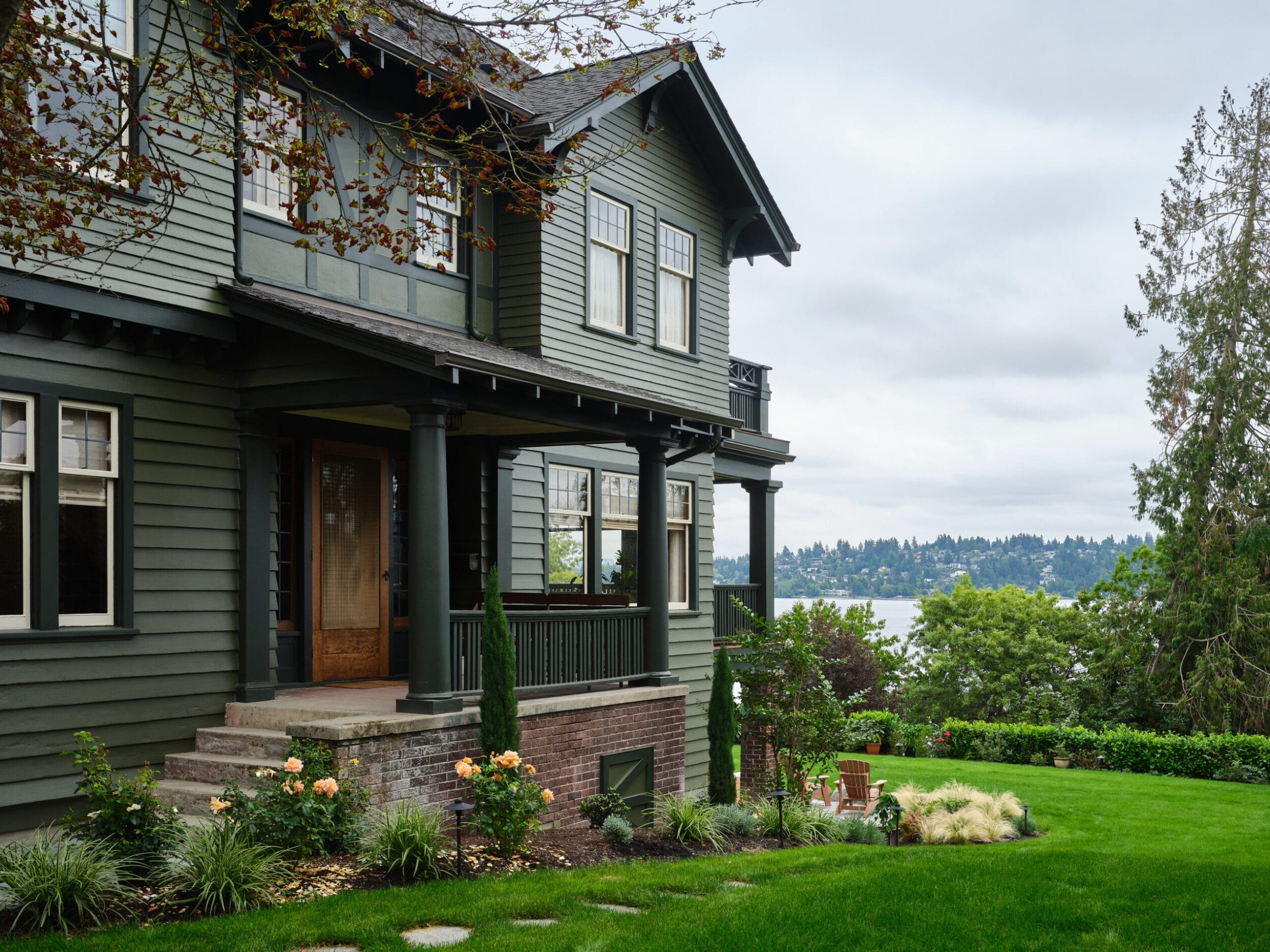 Green two-story house with a porch, surrounded by a lush garden and trees, overlooking a body of water in the background under a cloudy sky.