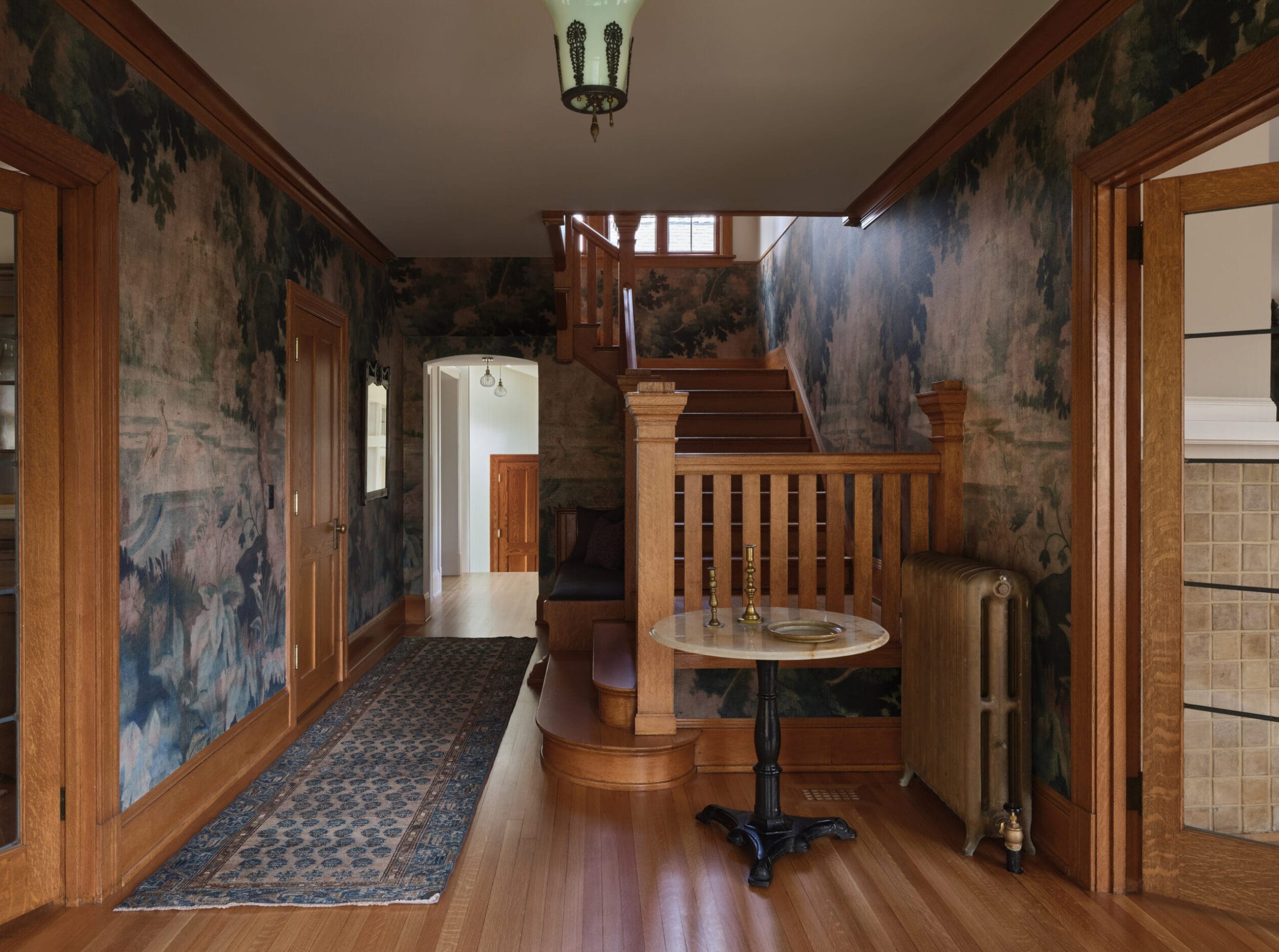 A hallway with wood flooring, a staircase, patterned wallpaper, a rug, a small round table with candlesticks, and a wooden radiator cover.