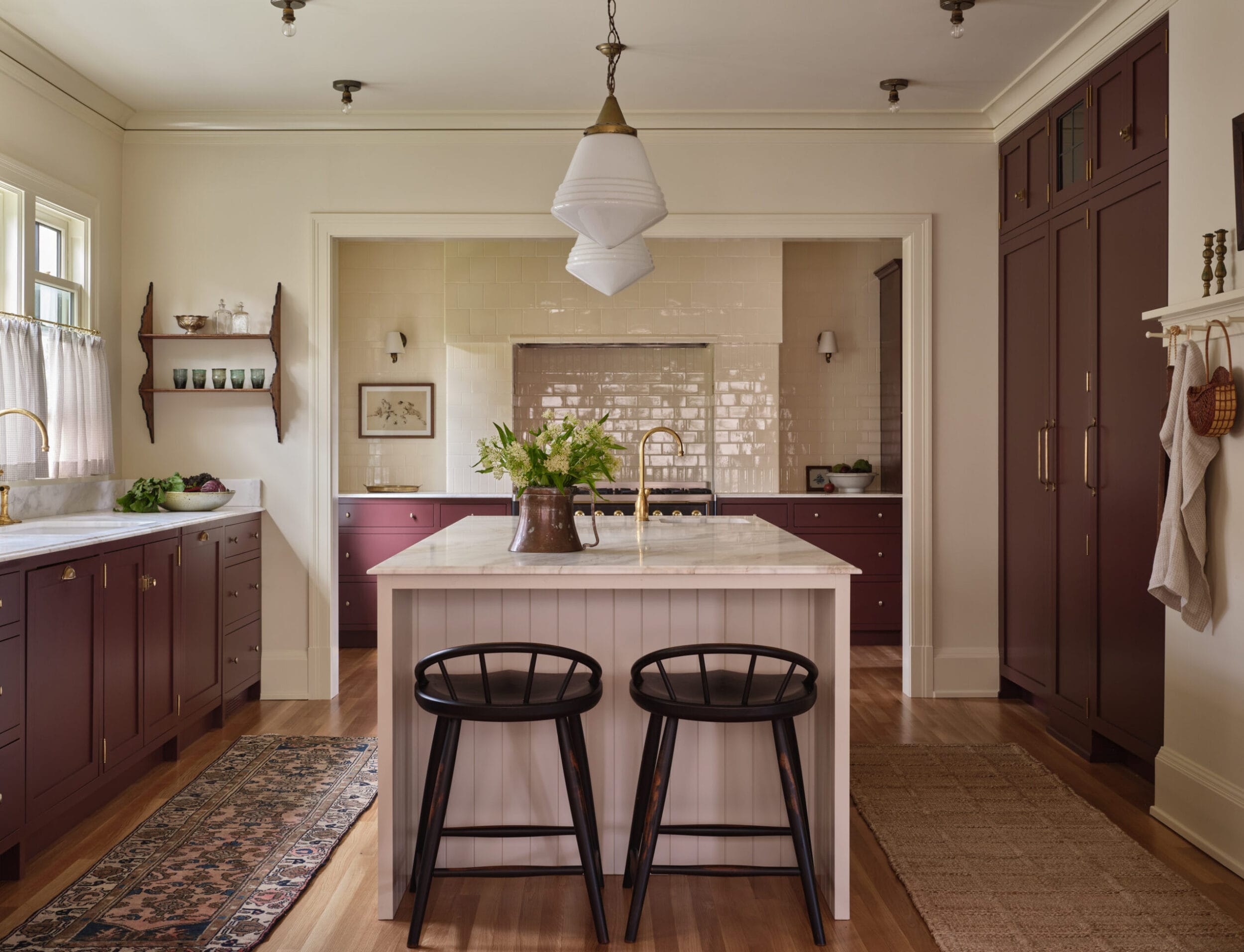 A kitchen with maroon cabinets, white island, wooden stools, and pendant light. A vase with flowers is on the island. There are windows, rugs, and a coat rack with hanging items.