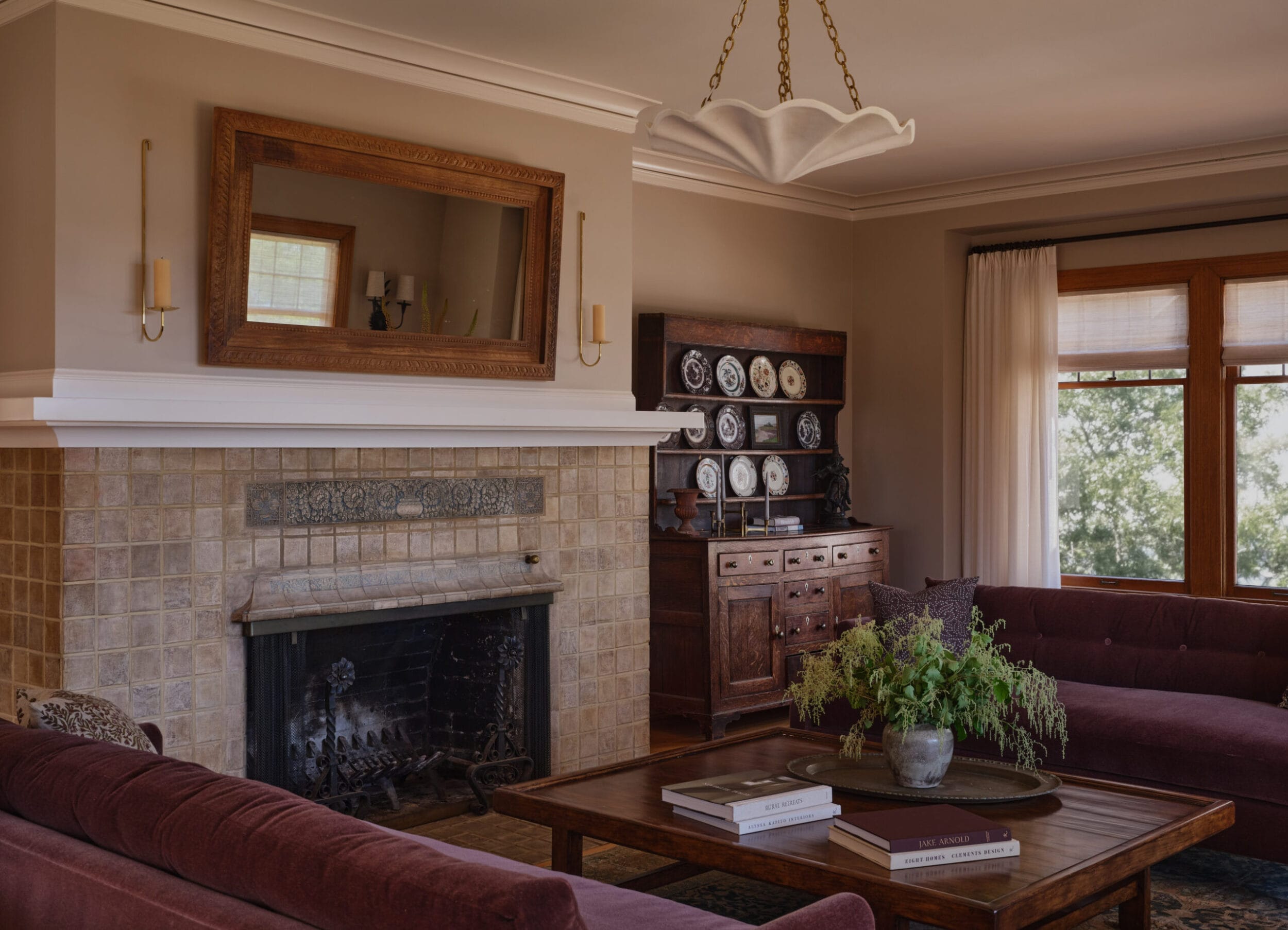 A living room with a tiled fireplace, a large mirror above it, two sofas, a wooden coffee table with a plant, and a cabinet displaying plates. A chandelier hangs from the ceiling.