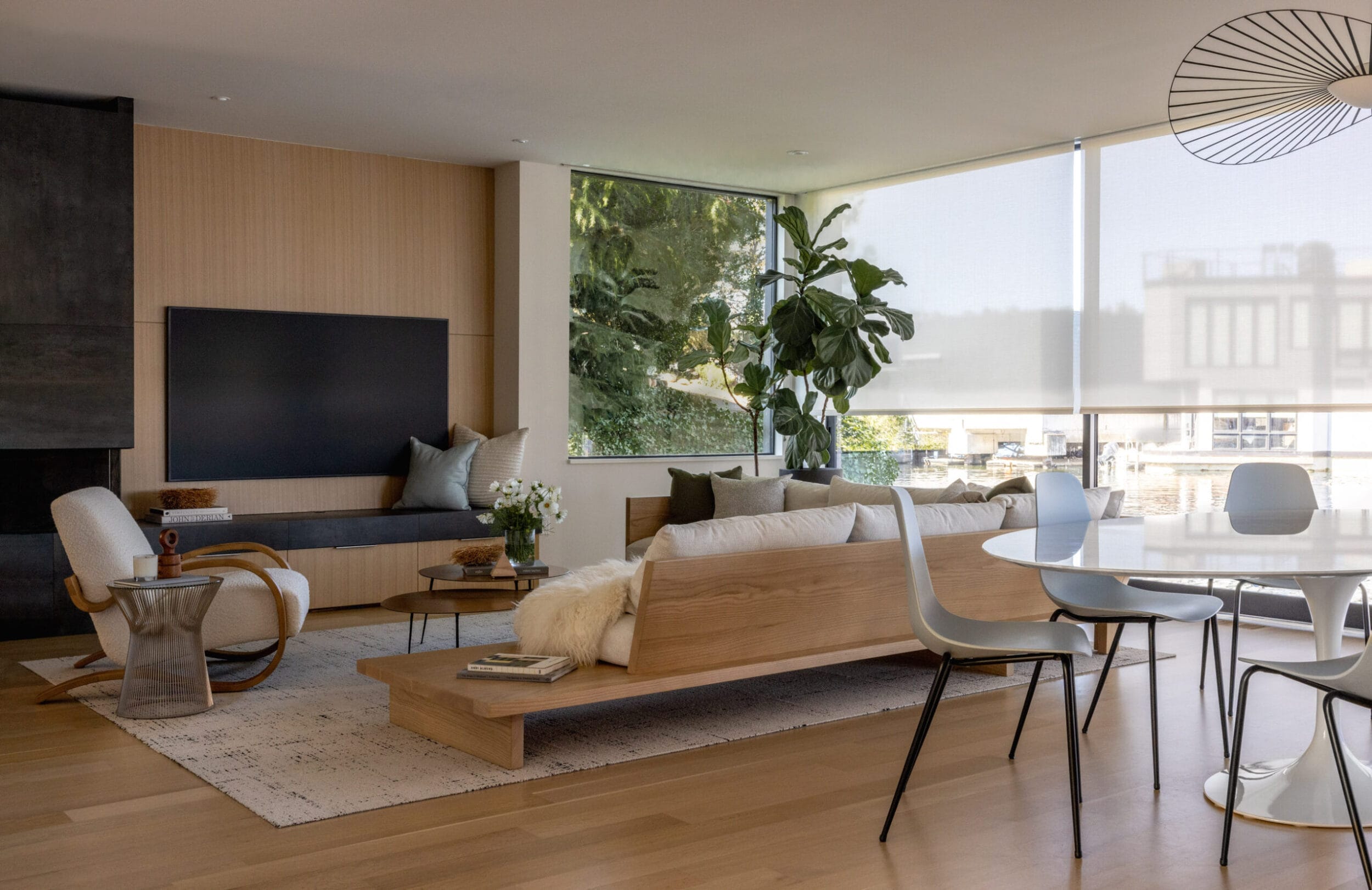 Modern living room with wooden floors, a low-profile couch, and a large plant by the window. Adjacent dining area features white chairs and a round table. Neutral and minimalist decor.