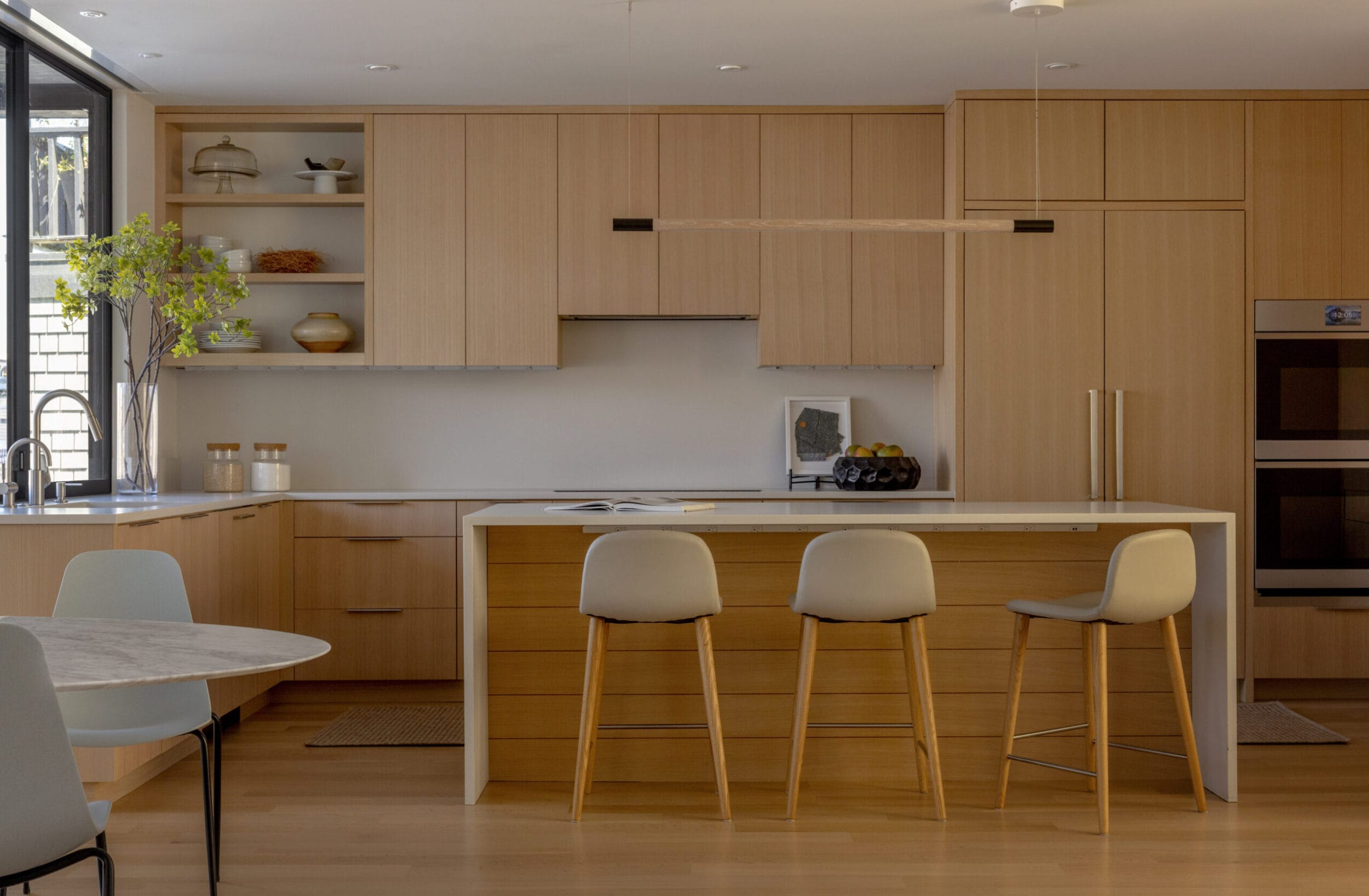 Modern kitchen with wooden cabinets, a white countertop island with three stools, built-in oven, and dining table. Large window provides natural light.