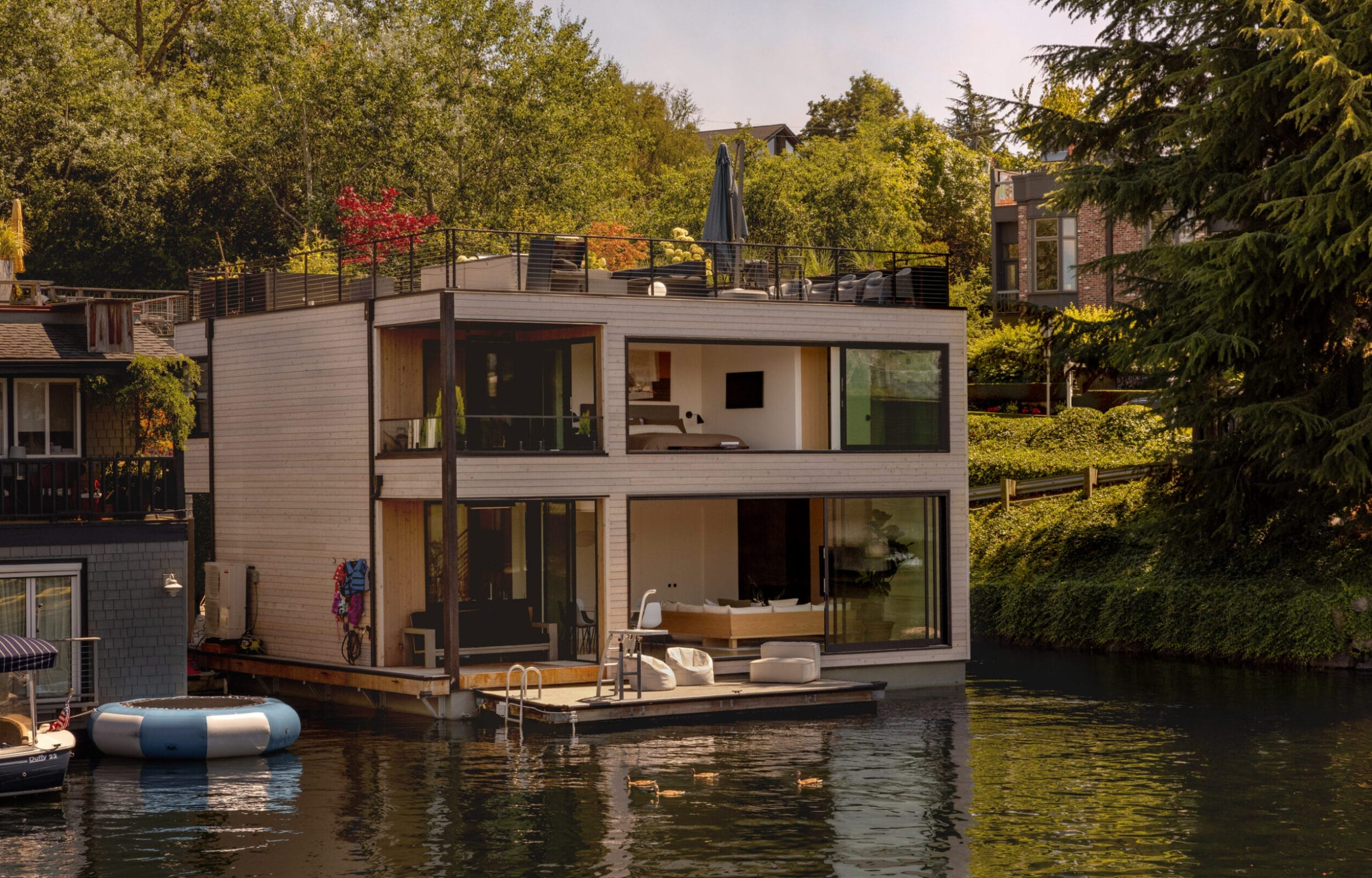 A modern two-story houseboat with large glass windows floats on a calm waterway, surrounded by trees and neighboring homes. There are outdoor seating and a raft nearby.