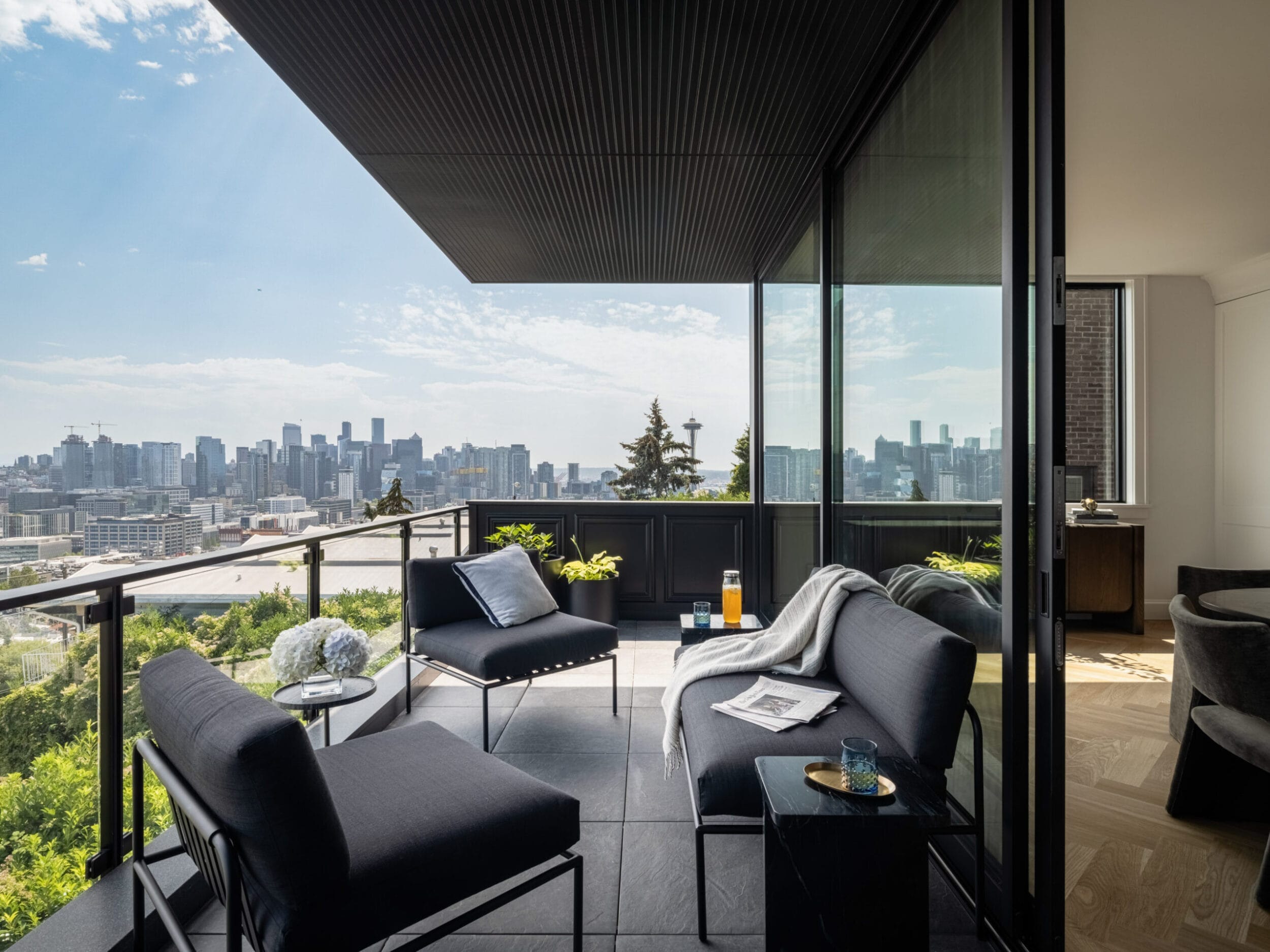 A modern balcony with black seating overlooks a city skyline. The space includes a table with drinks and a blanket. The sky is clear with some clouds.