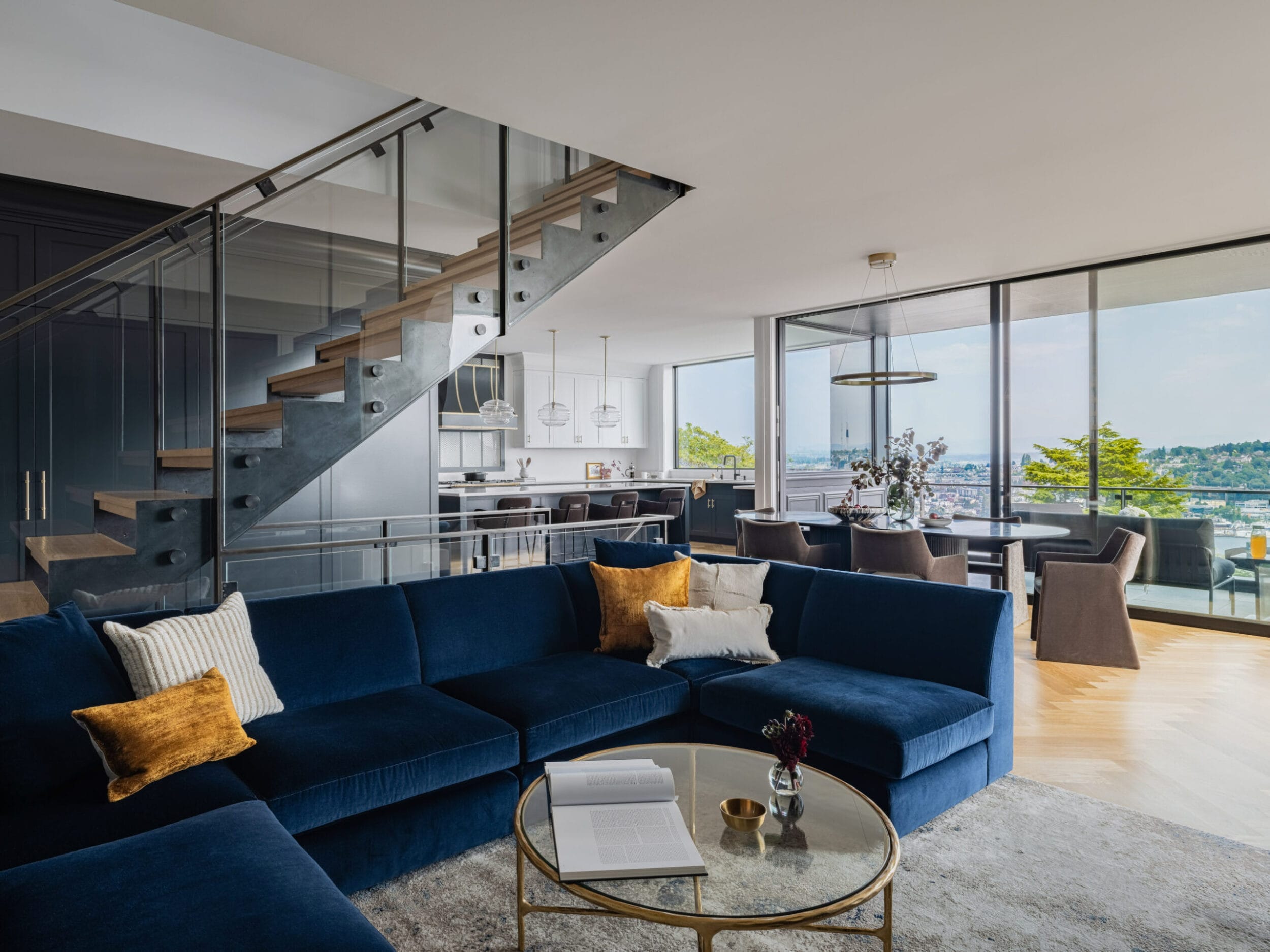 Modern living room with a blue sectional sofa, glass coffee table, and open wooden staircase. Large windows offer a view of a cityscape. Dining area with a table and chairs in the background.