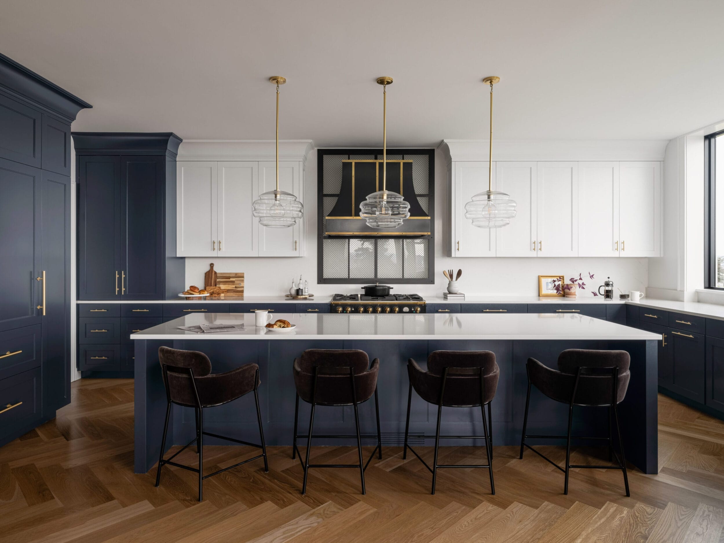 Modern kitchen with a large island, pendant lights, navy and white cabinets, bar stools, and a herringbone wood floor.