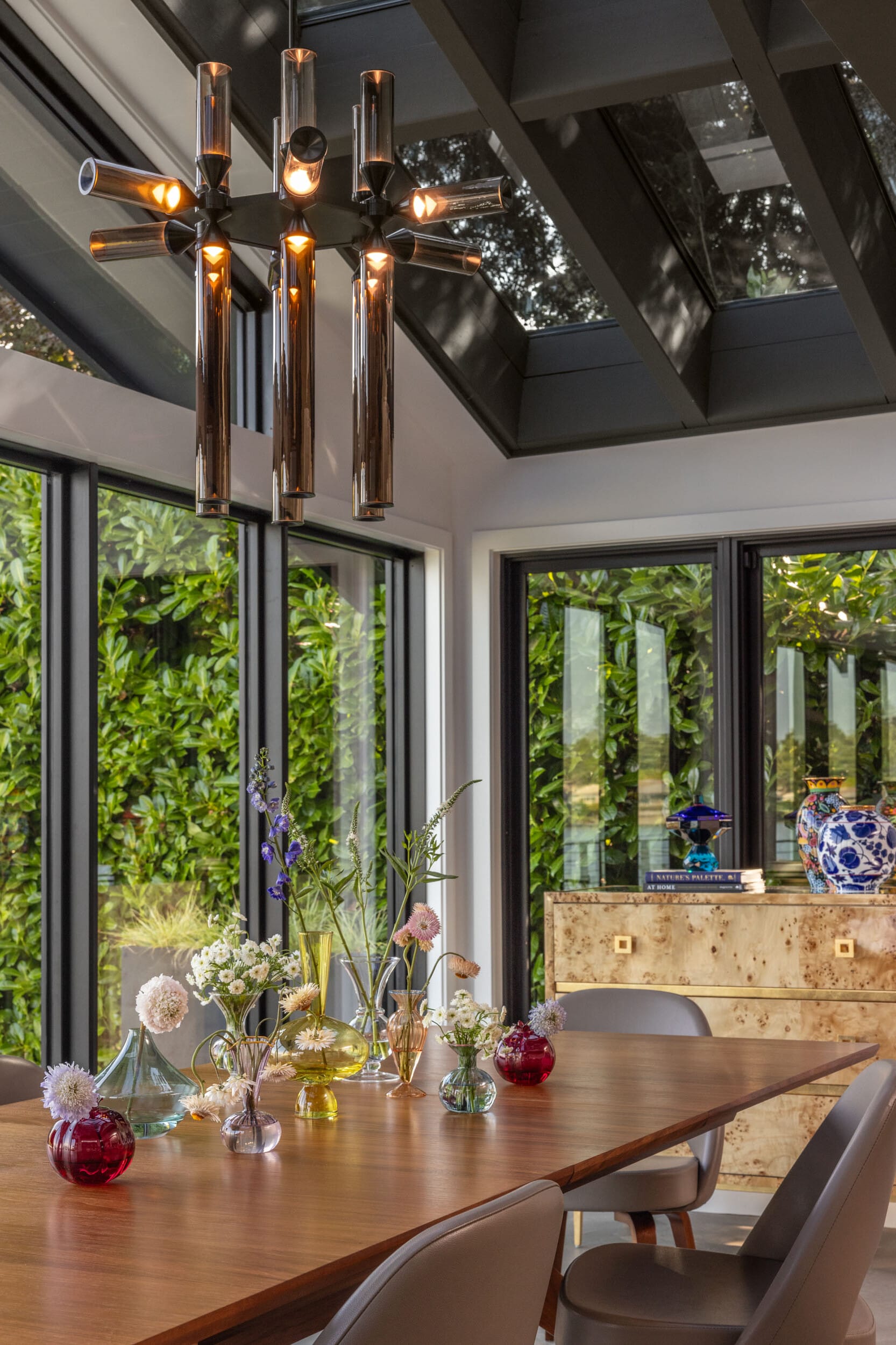 A dining room with a wooden table, modern chandelier, colorful vases, and a sideboard. Large windows reveal greenery outside.