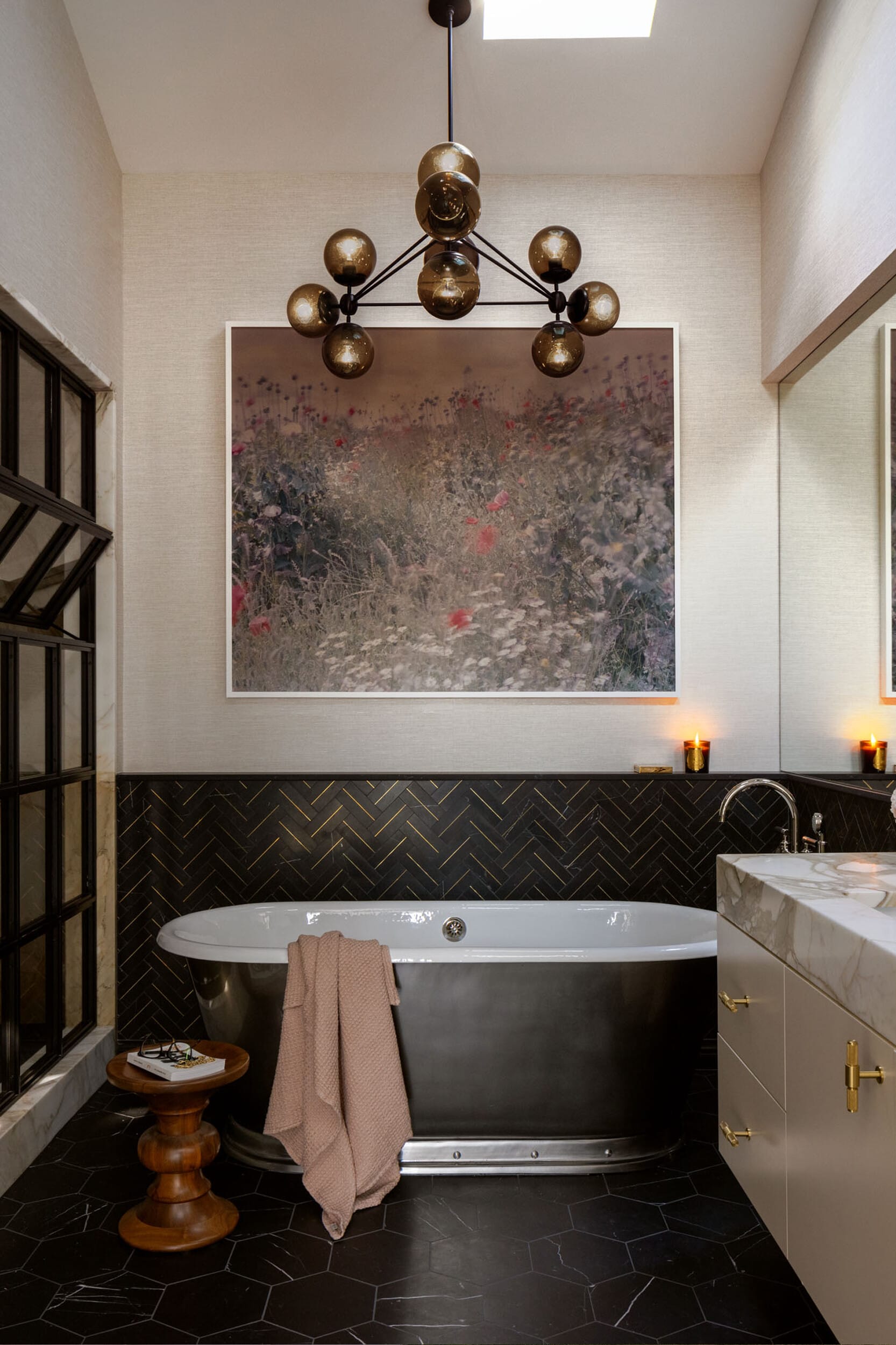 Elegant bathroom with a black freestanding tub, herringbone tiling, large floral artwork, modern chandelier, and a marble-topped vanity. A folded towel drapes over a wooden stool.