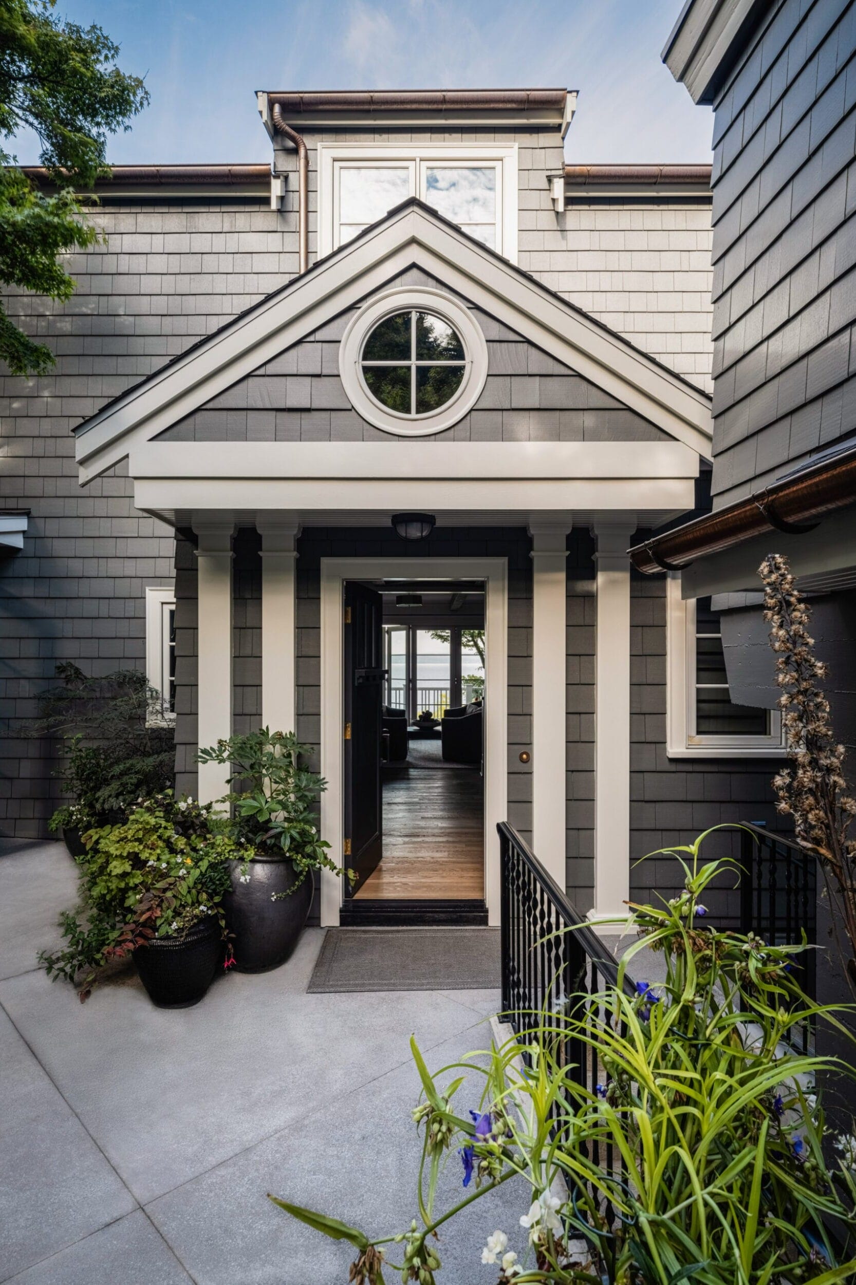 Front view of a gray shingle-style house with a round window, white trim, and an open front door. The entrance is flanked by potted plants and framed by a peaked roof.