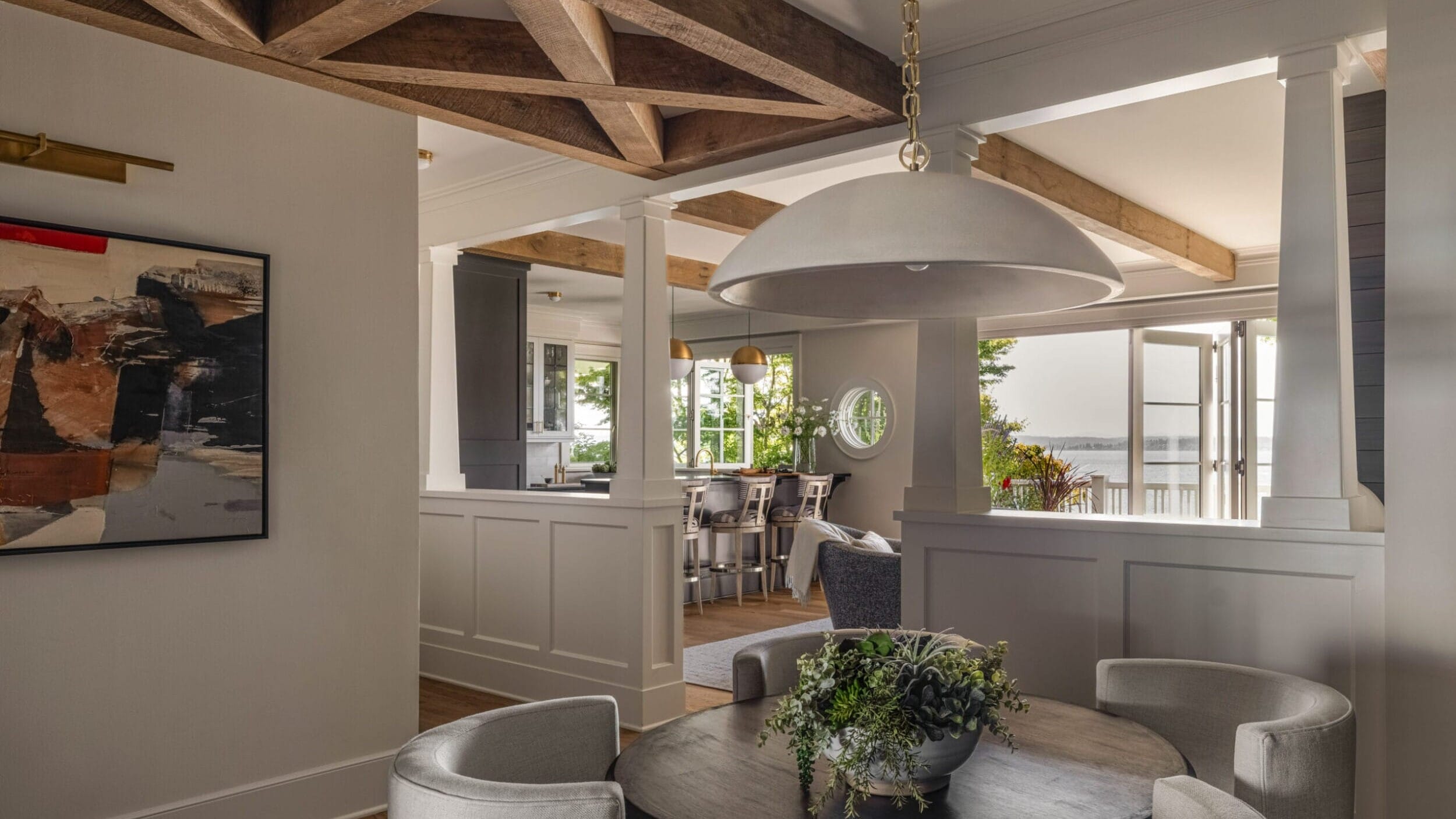 Modern dining room with a round table and plush chairs, featuring exposed wooden beams and a large pendant light. An open doorway leads to a brighter area with windows overlooking a body of water.