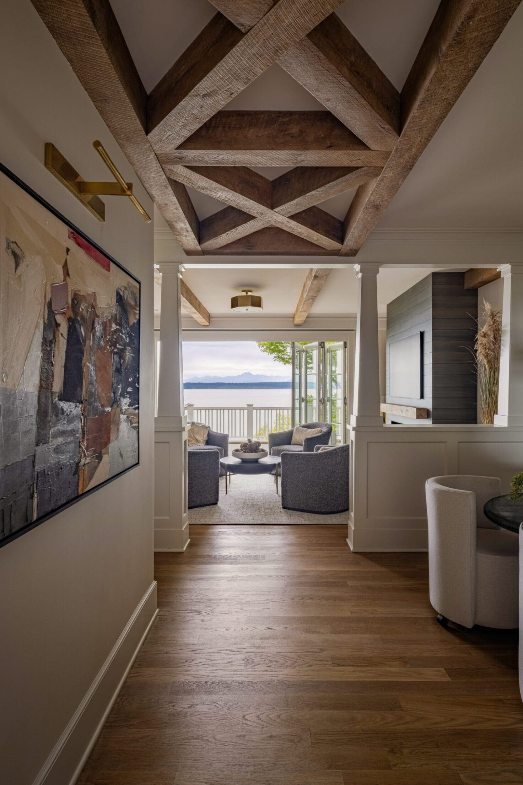 View of a hallway leading to a seating area with armchairs and a large window overlooking a body of water, featuring exposed wooden ceiling beams and abstract wall art.