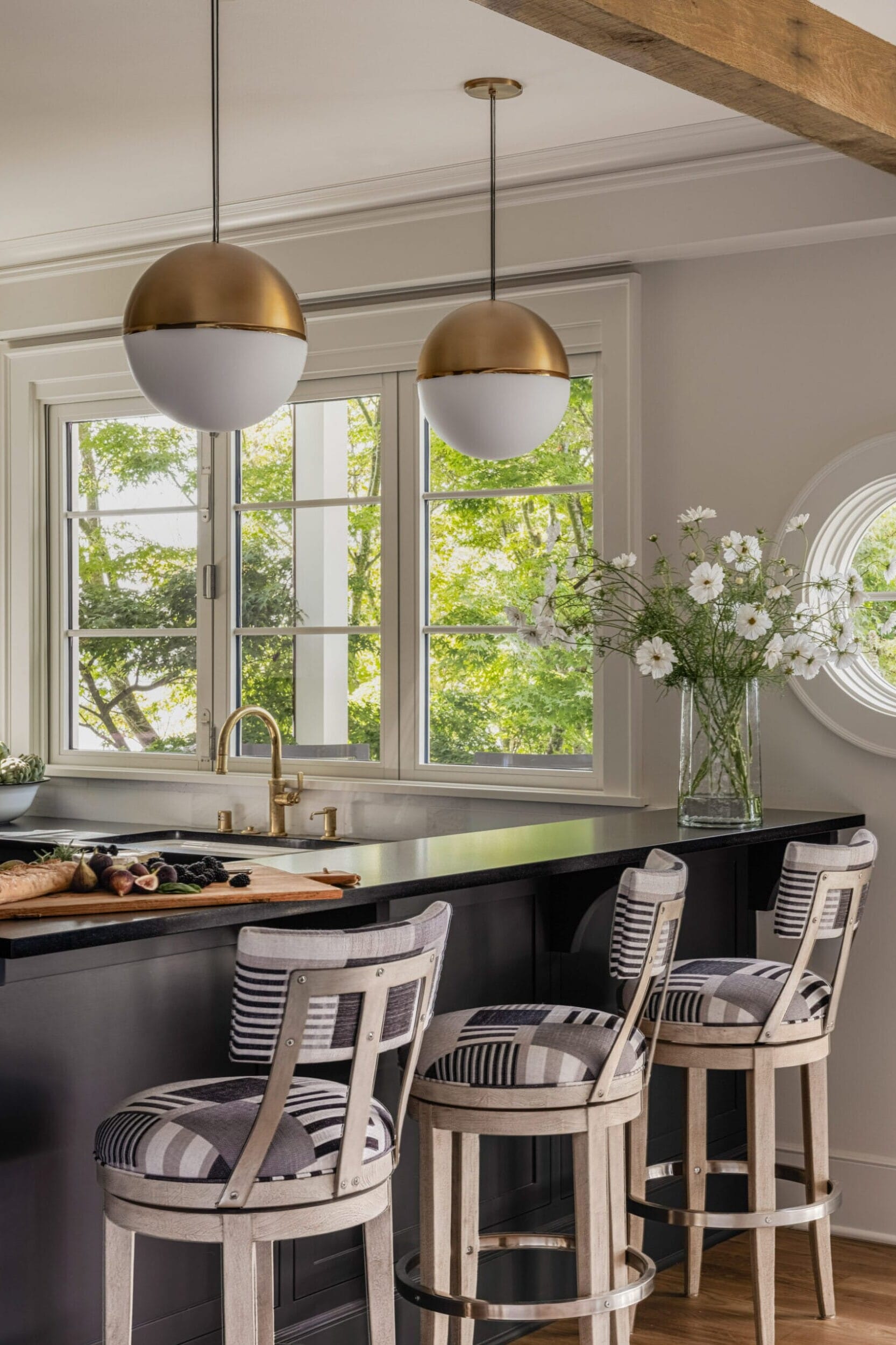 A kitchen with three striped stools at a dark counter, pendant lights, round window, and a vase of white flowers. Large windows show greenery outside.