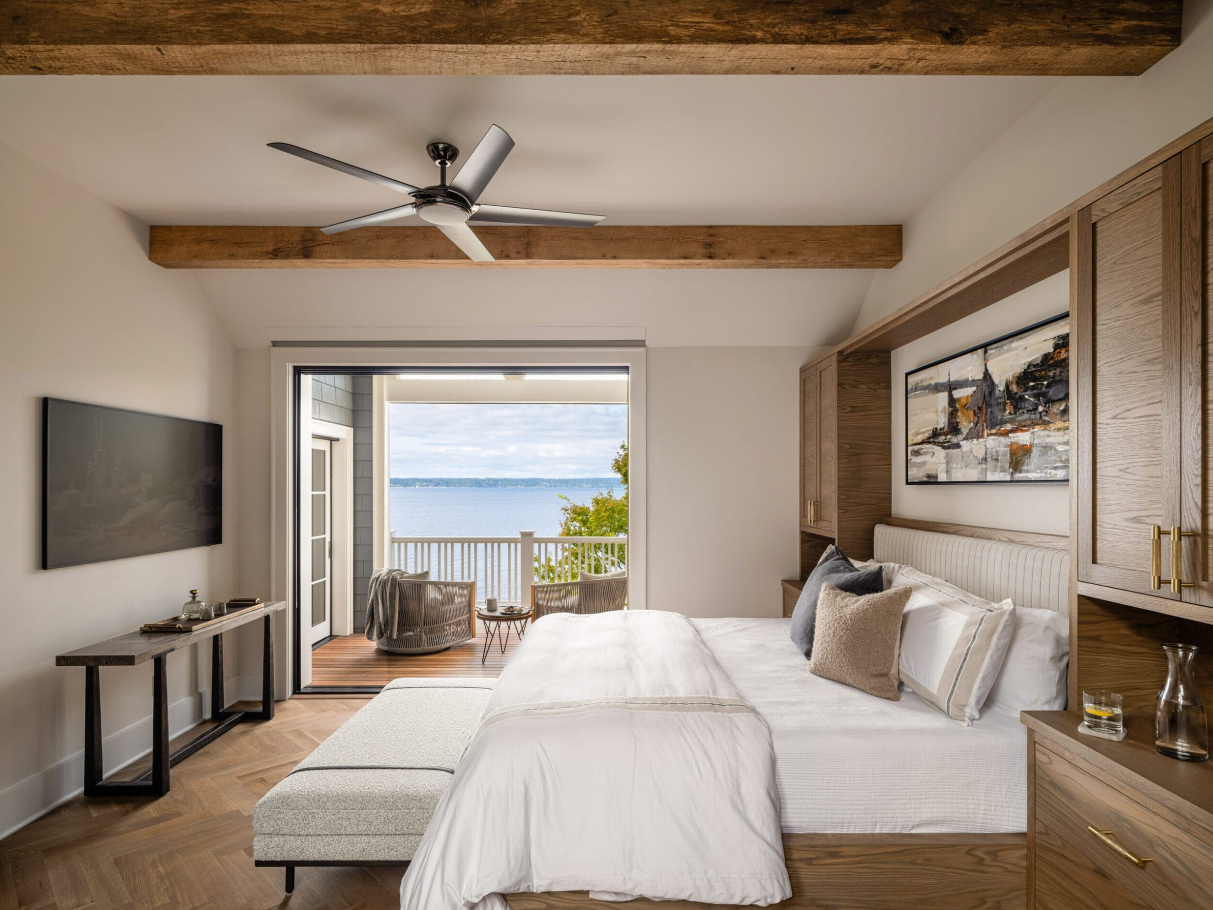 A bedroom with a white bed, wooden beams, and a ceiling fan. It opens to a balcony with chairs and a view of a large body of water and trees. A TV and console are on the left wall.