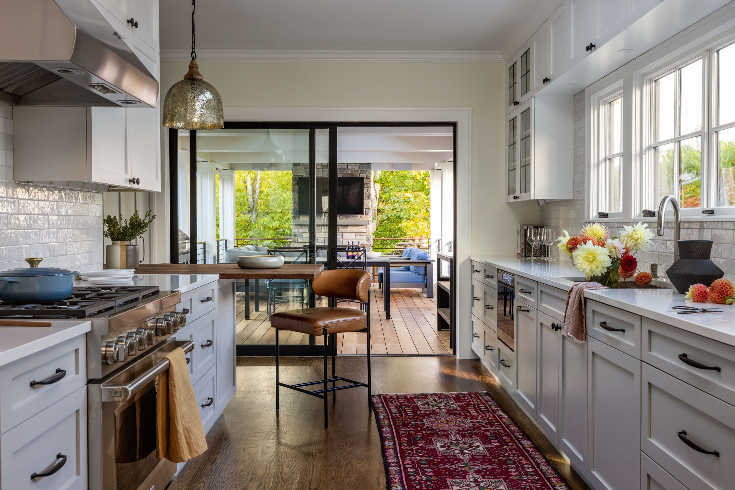 Modern kitchen with white cabinets, a central wooden island with a bar stool, and a patterned rug. Sliding glass doors open to a deck with seating. Bright flowers are displayed on the countertop.