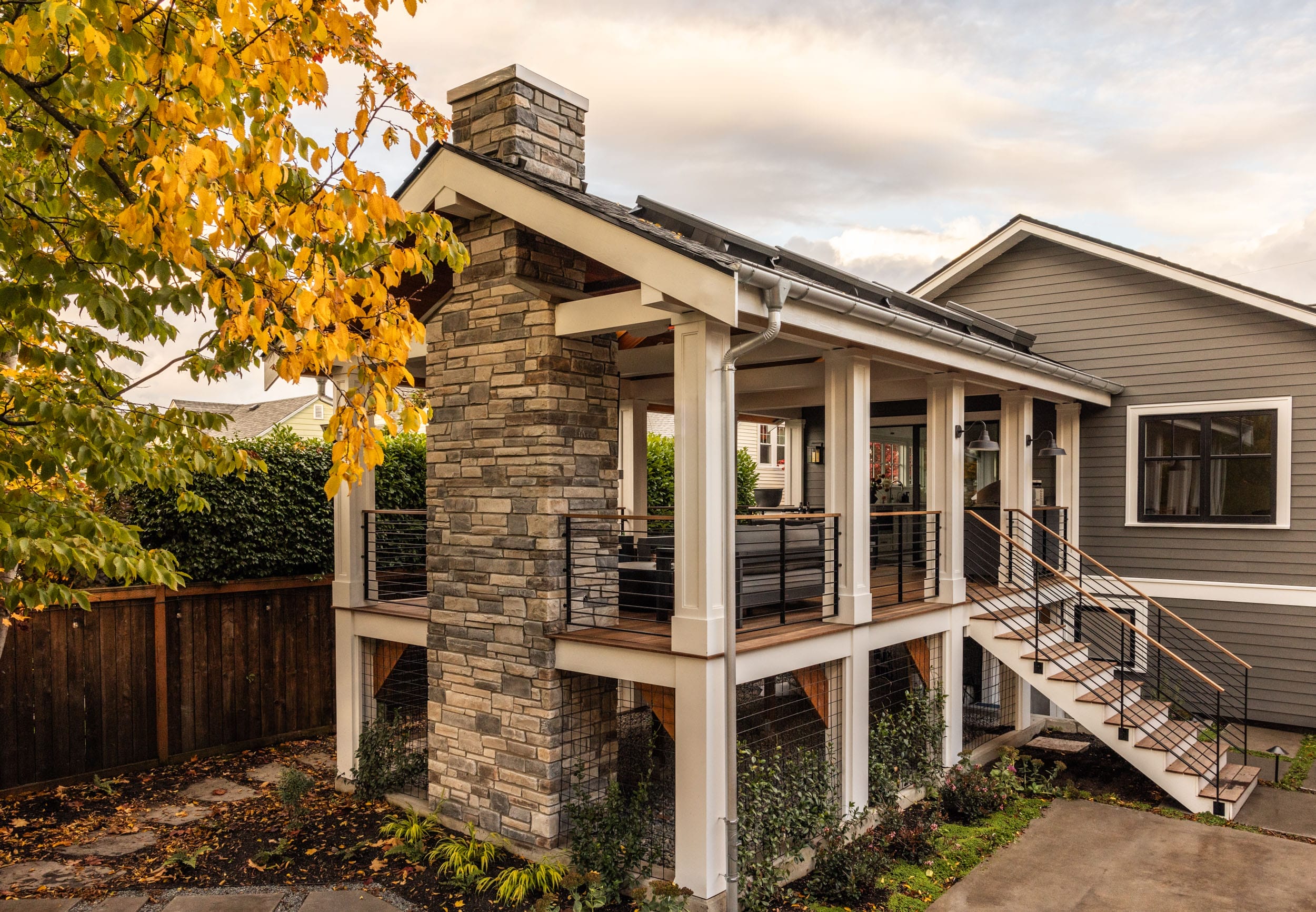 A two-story house with a stone chimney, covered porch, and outdoor staircase. Surrounded by trees and a wooden fence.