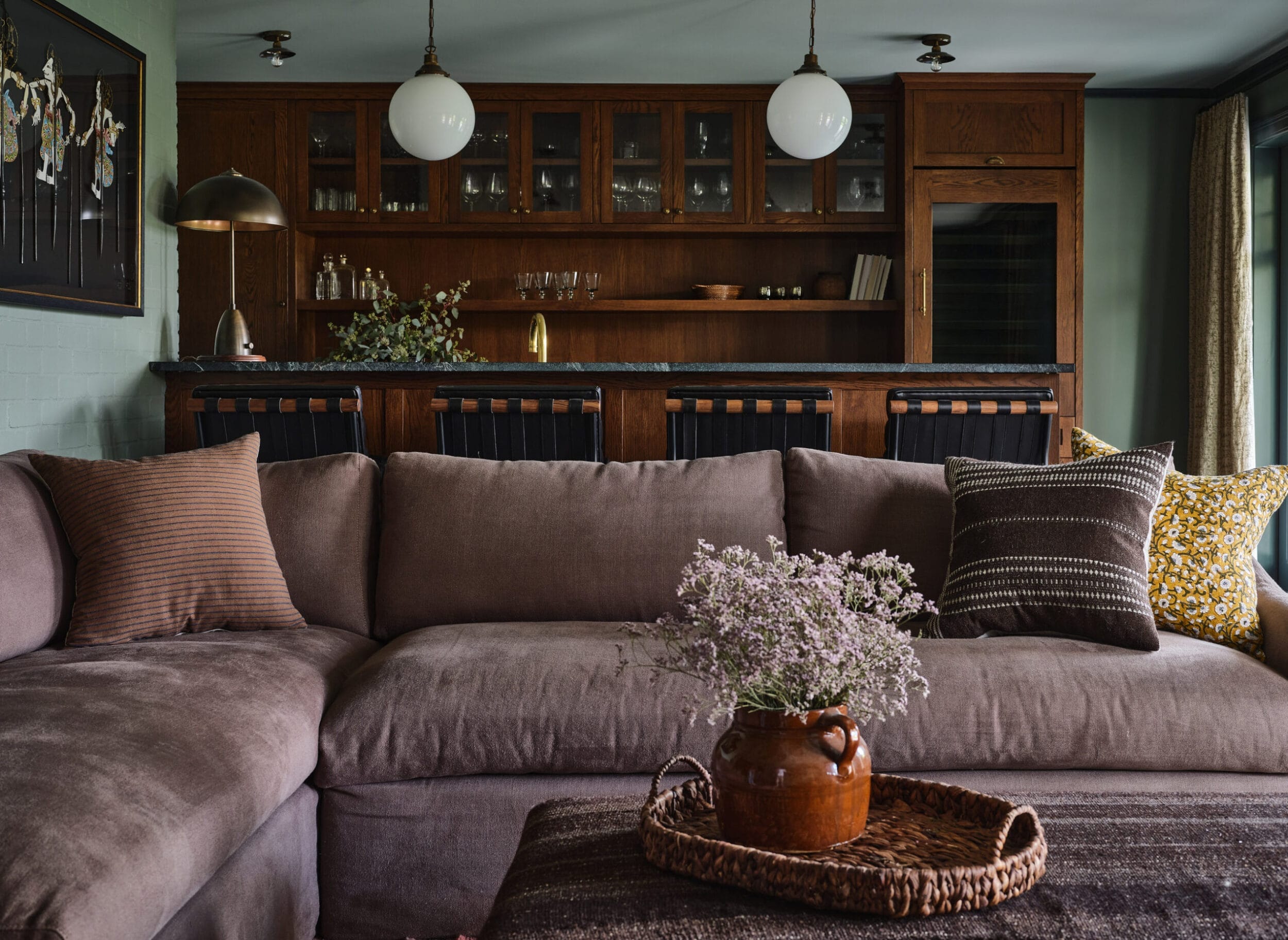 A cozy living room with a large gray sectional sofa, decorative pillows, and a woven tray with flowers. In the background, there's a wooden bar with glass cabinets and two hanging lights.