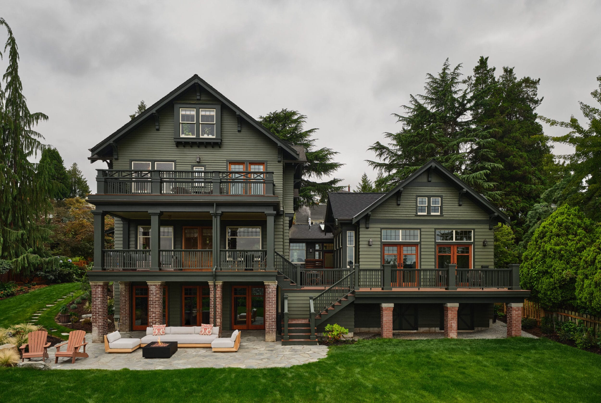 A large two-story house with green siding and multiple balconies is surrounded by trees and a manicured lawn. Outdoor seating is arranged on a stone patio in the backyard.