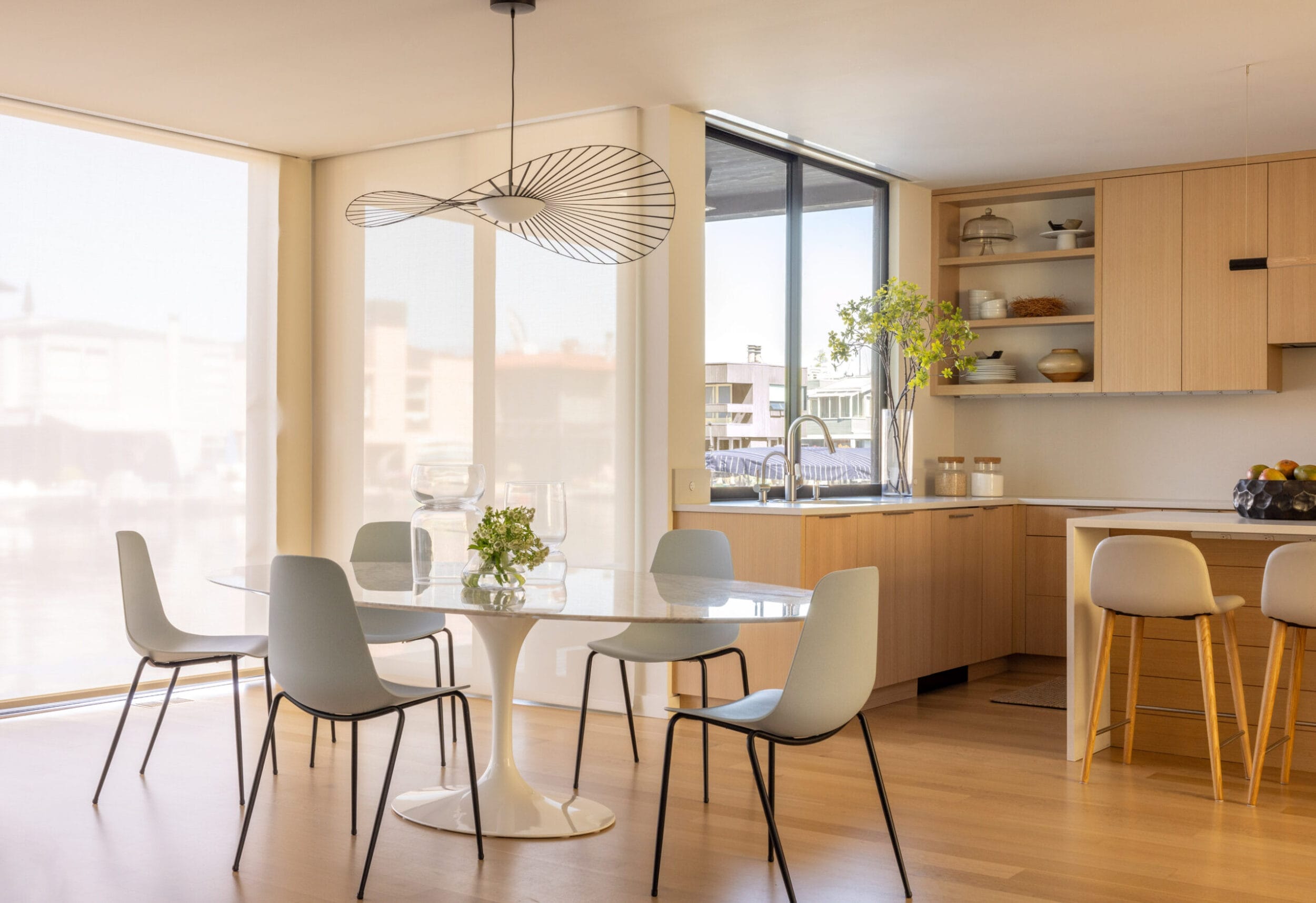 A modern kitchen and dining area with light wood cabinets, a marble table with black chairs, a large window, and a unique overhead light fixture.