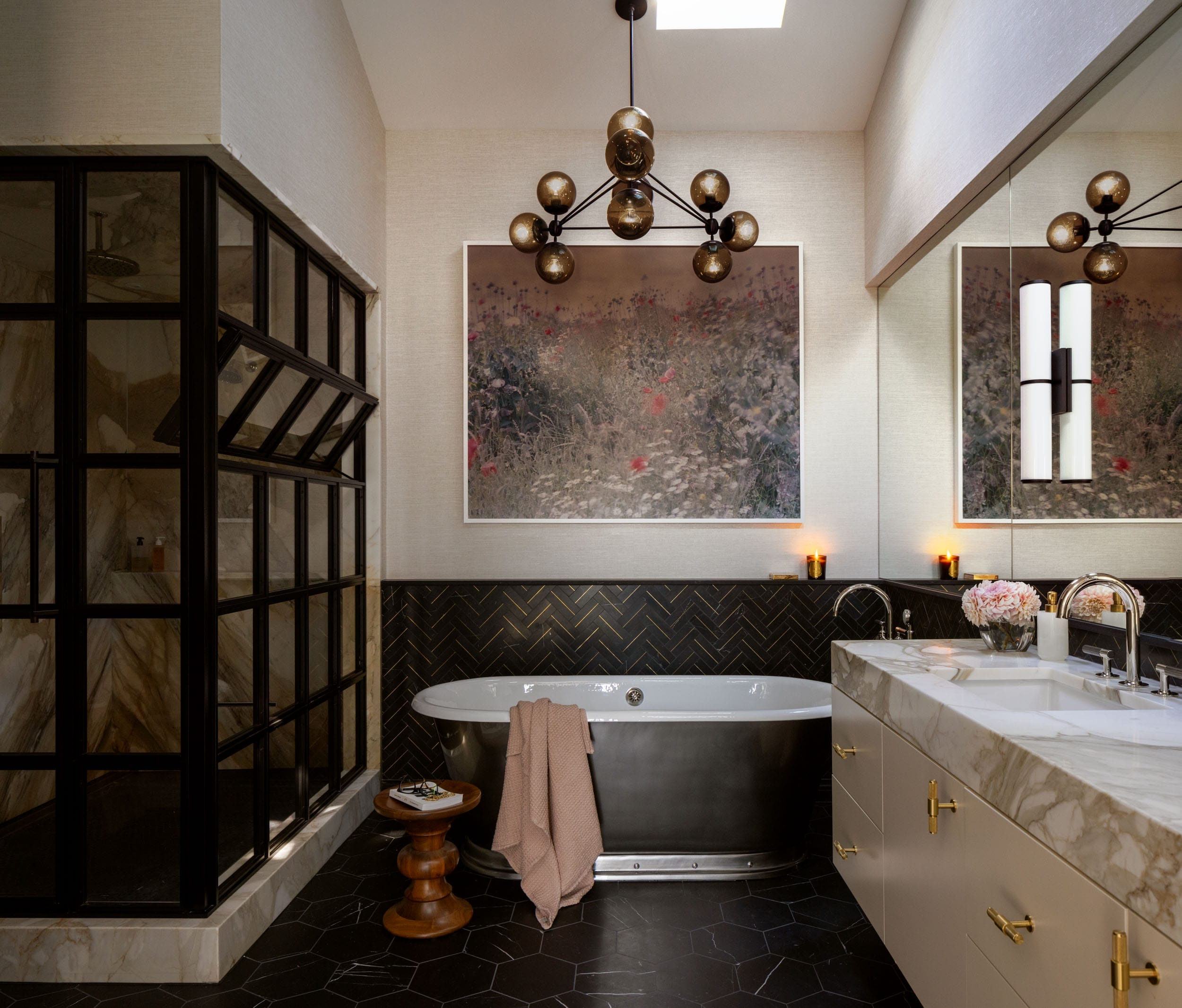 A luxurious bathroom with a dark free-standing bathtub, modern chandelier, large mirror, and black hexagonal floor tiles. A floral painting hangs above the bathtub.