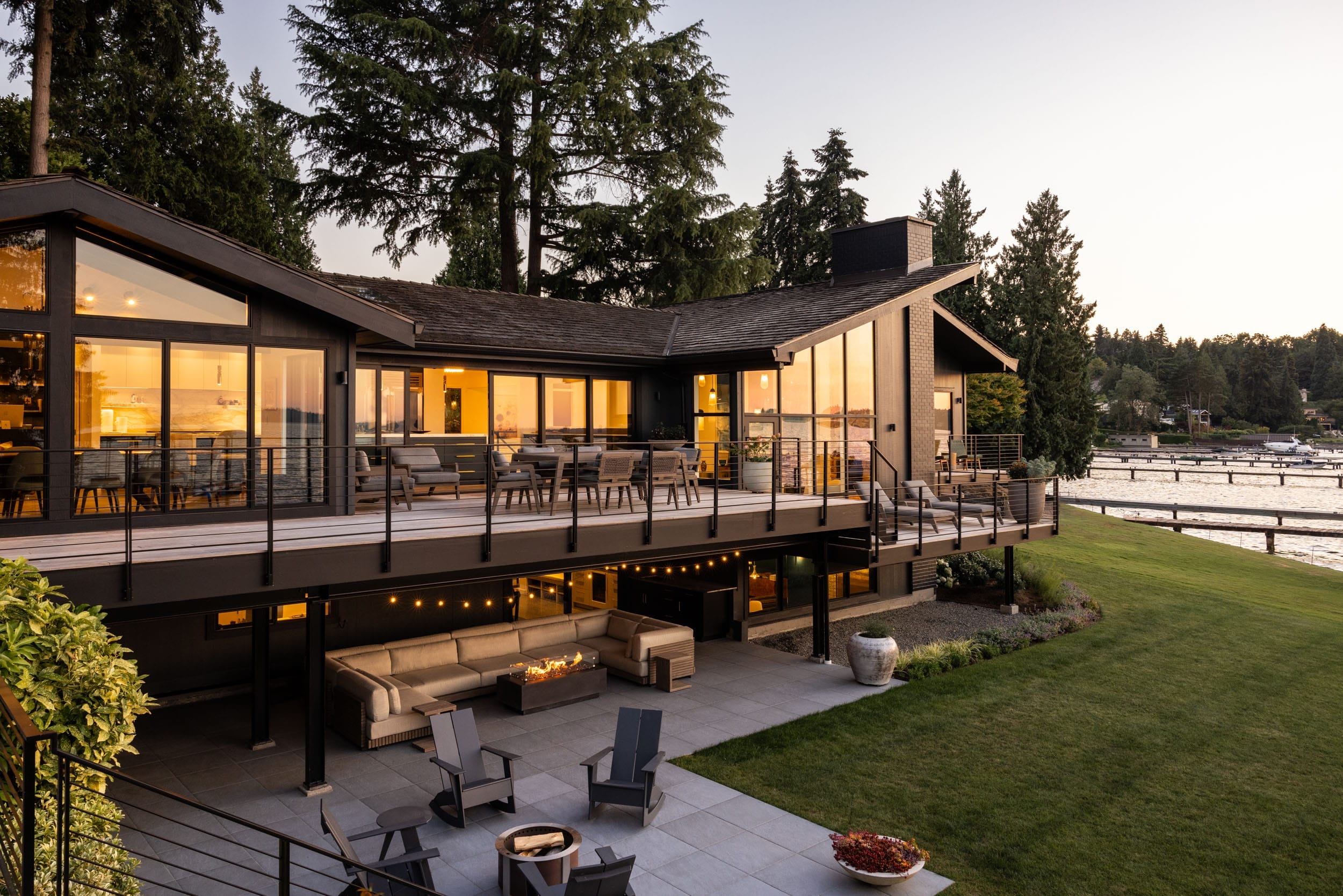 A modern two-story lakeside house with large windows, a spacious deck, outdoor seating, and a green lawn, surrounded by tall trees at sunset.