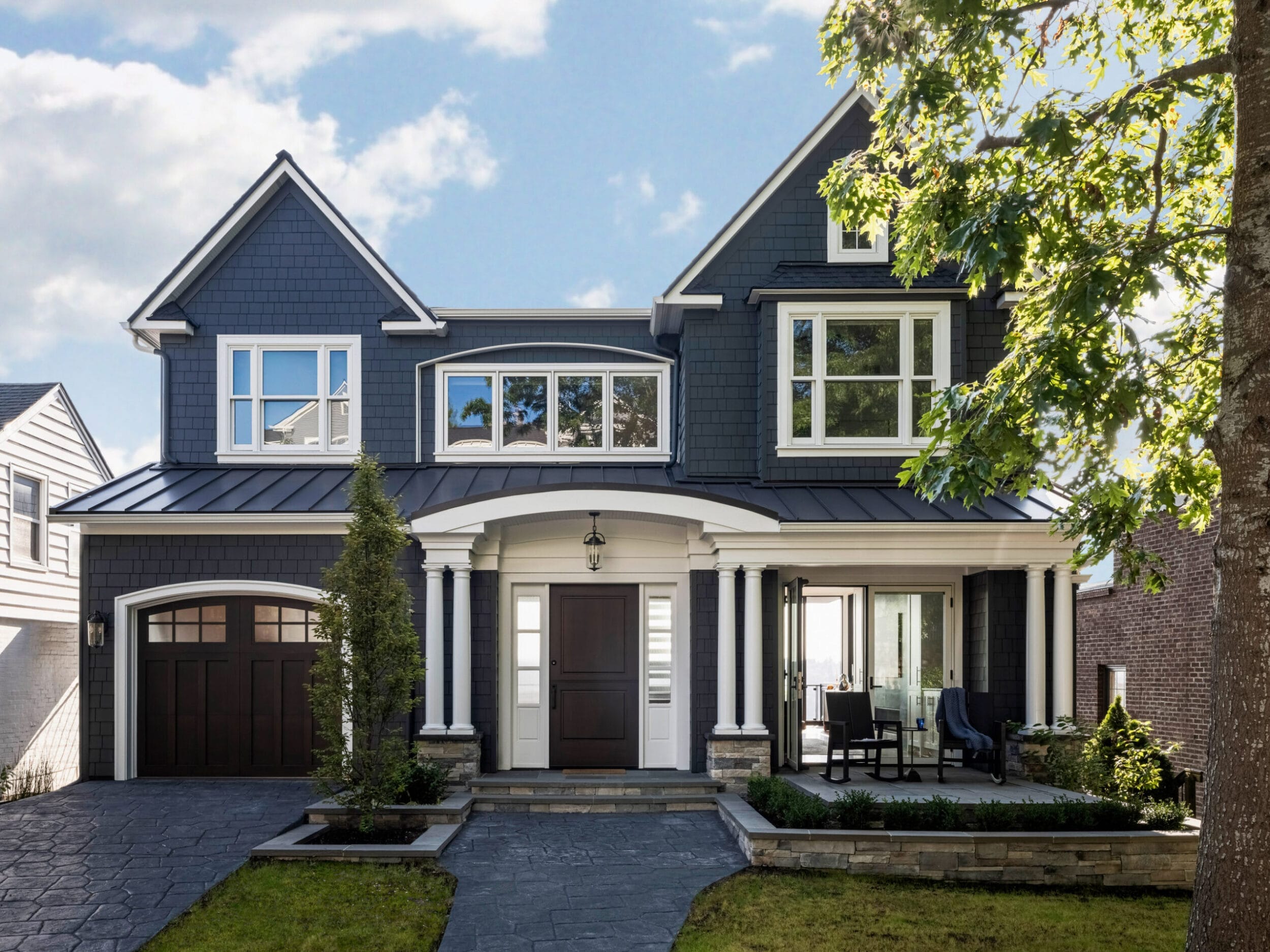 Two-story house with dark gray siding, white trim, and a covered front porch. There's a single garage door on the left and a small outdoor seating area on the right.