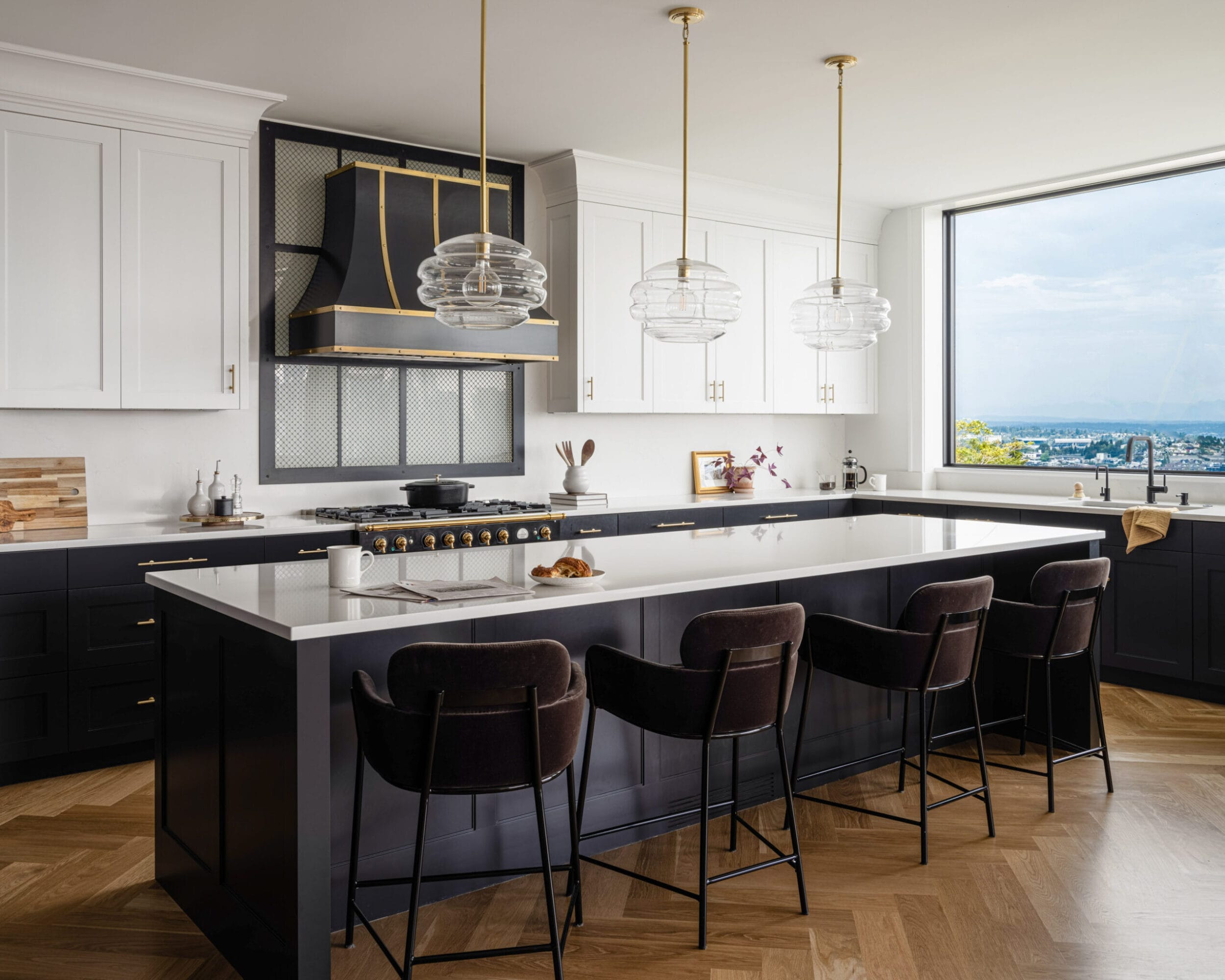 Modern kitchen with a large island, black and white cabinetry, gold accents, and three pendant lights. A large window offers a view of a cityscape.